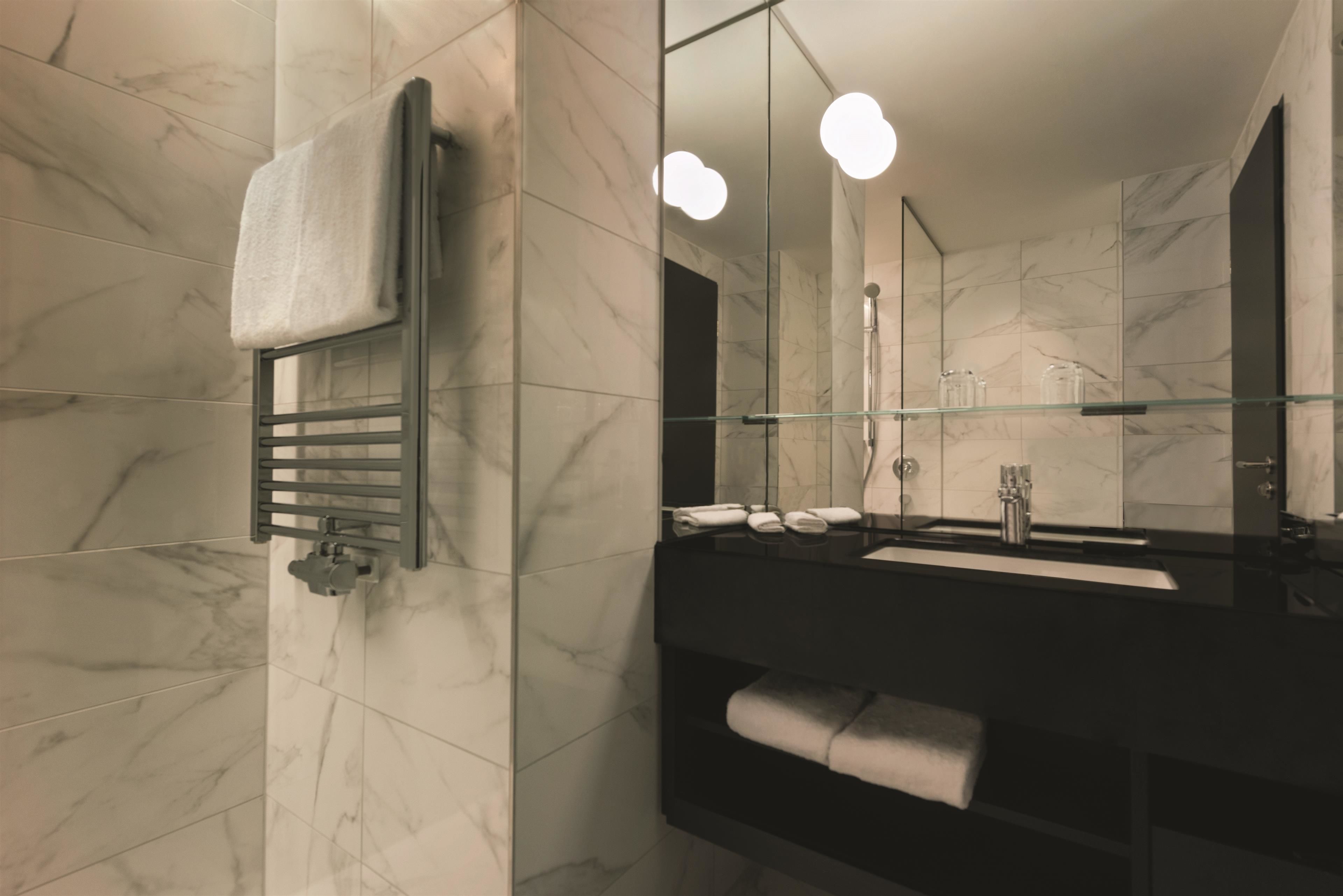 Un baño moderno con baldosas de mármol, cabina de ducha de cristal, lavabo negro y un espejo grande con luces.