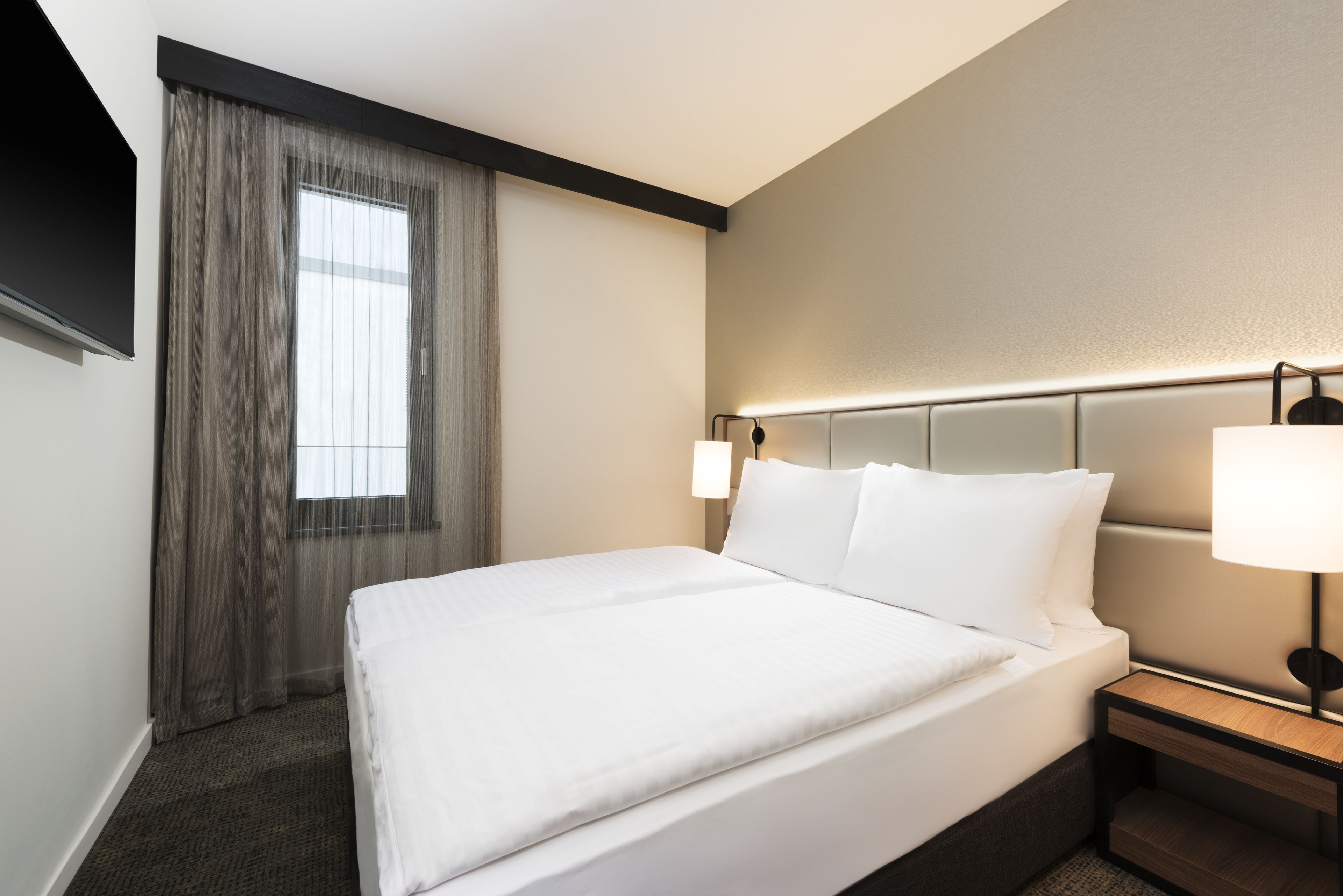 A hotel room with a large bed, a nightstand, a lamp, a window with curtains, and a wall-mounted TV.