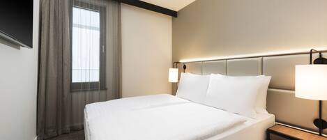 Habitación de hotel con una cama grande, una mesita de noche, una lámpara, una ventana con cortinas y un televisor montado en la pared.