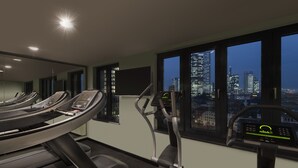A gym with treadmills and elliptical machines, large windows with a city skyline view, and a mirror reflecting the equipment. - Adina Apartment Hotel Frankfurt Westend (Frankfurt)