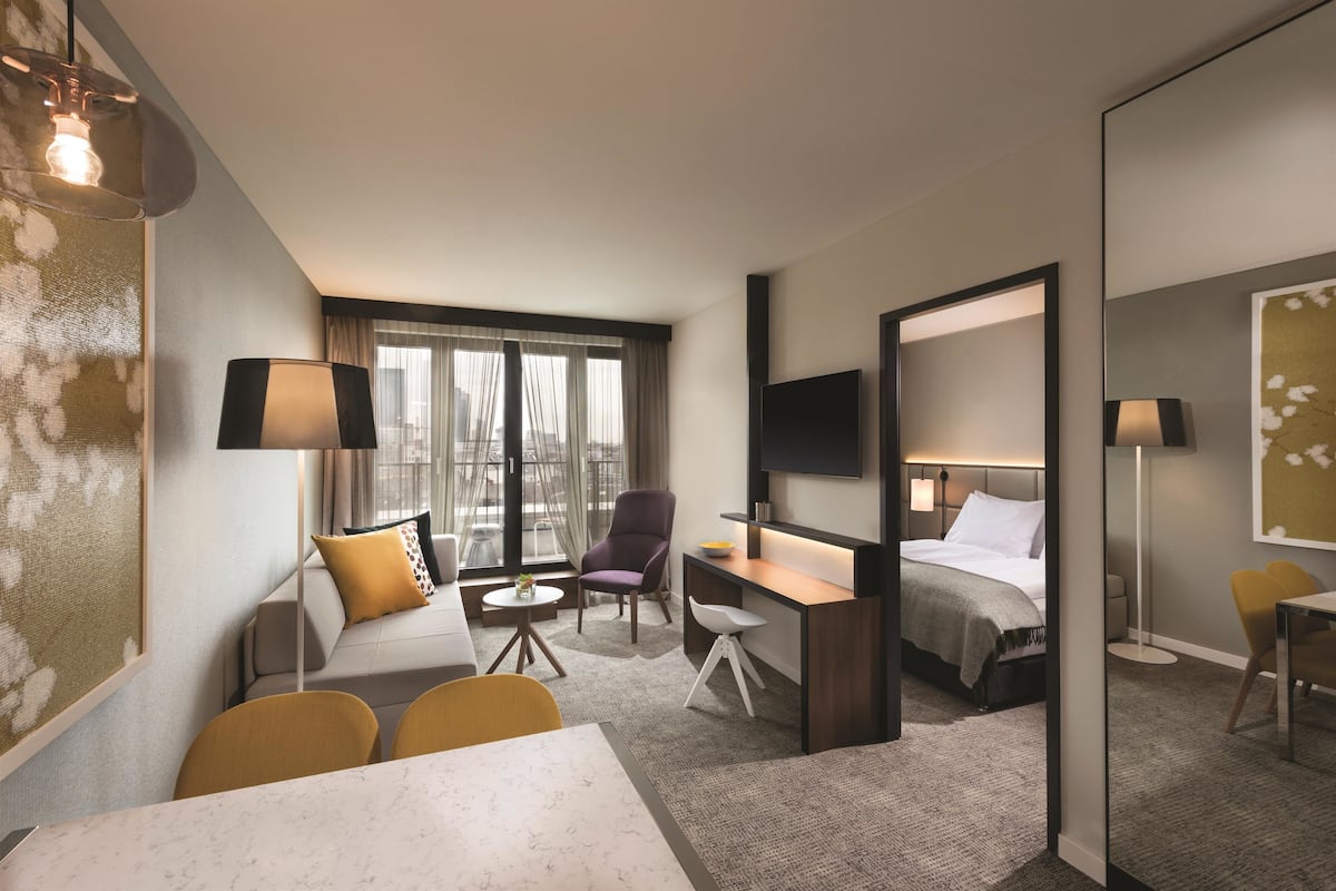 A hotel room with a sofa, armchair, desk, and a bed. There is a large window with curtains, a flat-screen TV mounted on the wall, and a small table with chairs.