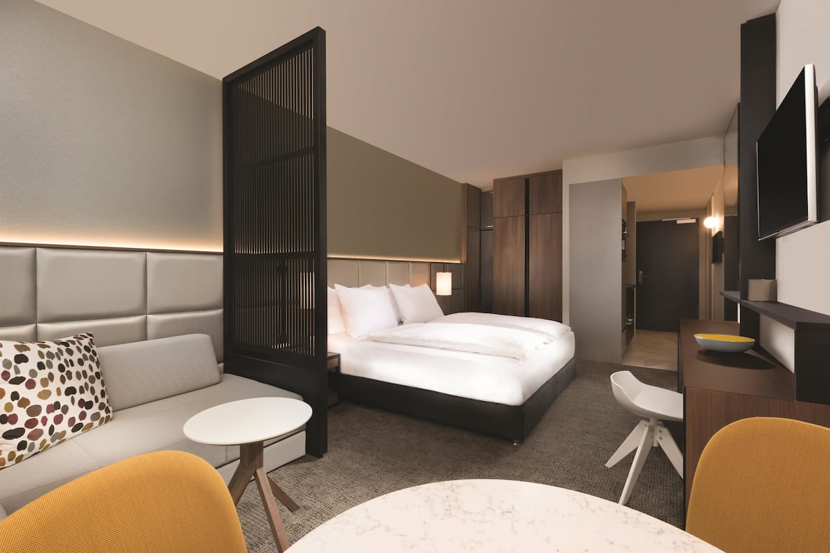 A modern hotel room with a bed, a sofa, a small round table, and a desk with a chair.