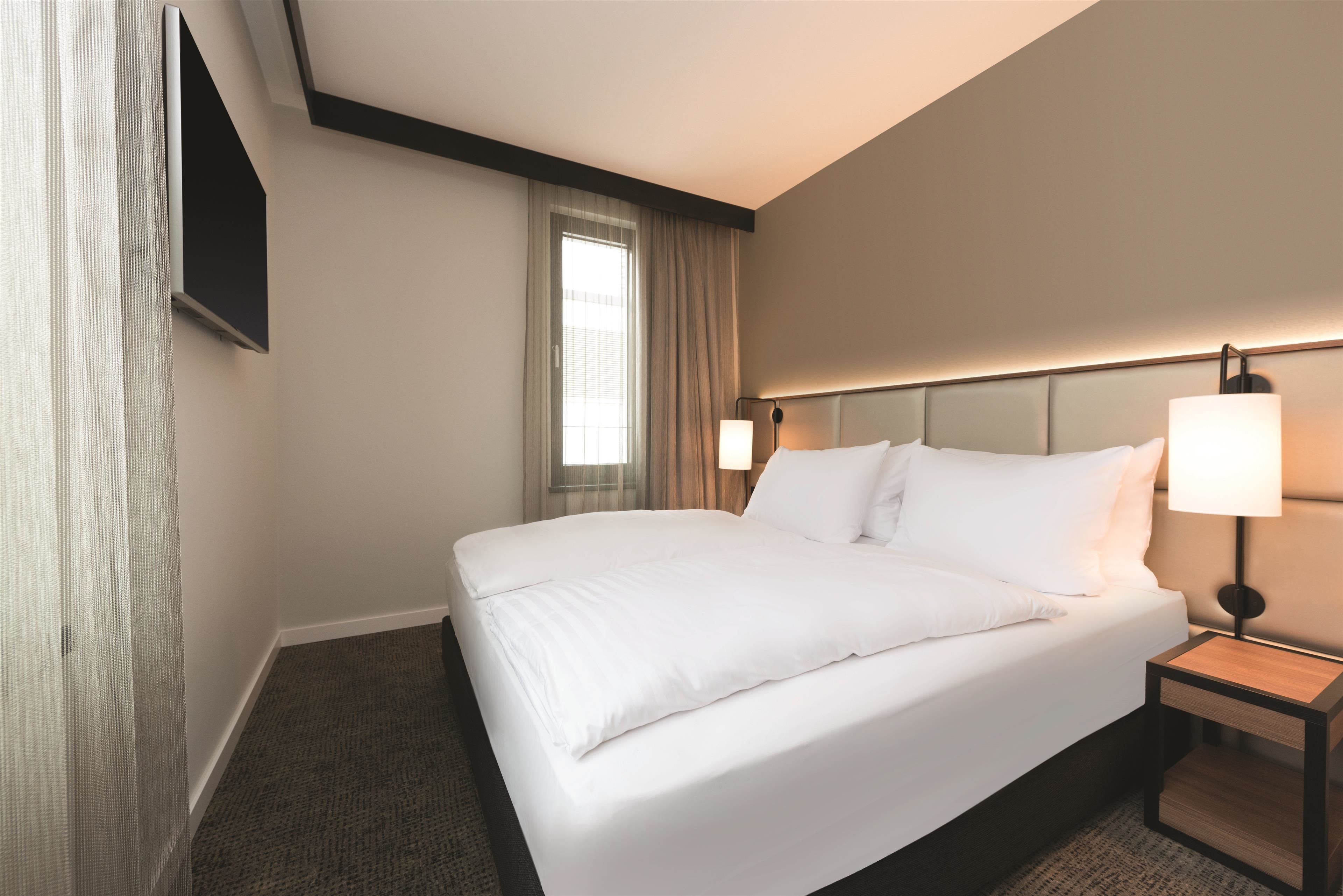 A hotel room with a large bed, a bedside table with a lamp, a window with curtains, and a flat-screen TV mounted on the wall.