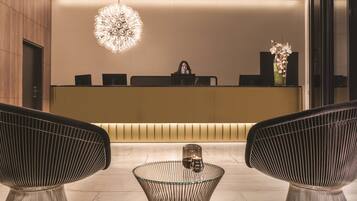 A modern reception area with a central desk, two upholstered chairs, a glass coffee table, and a decorative chandelier.