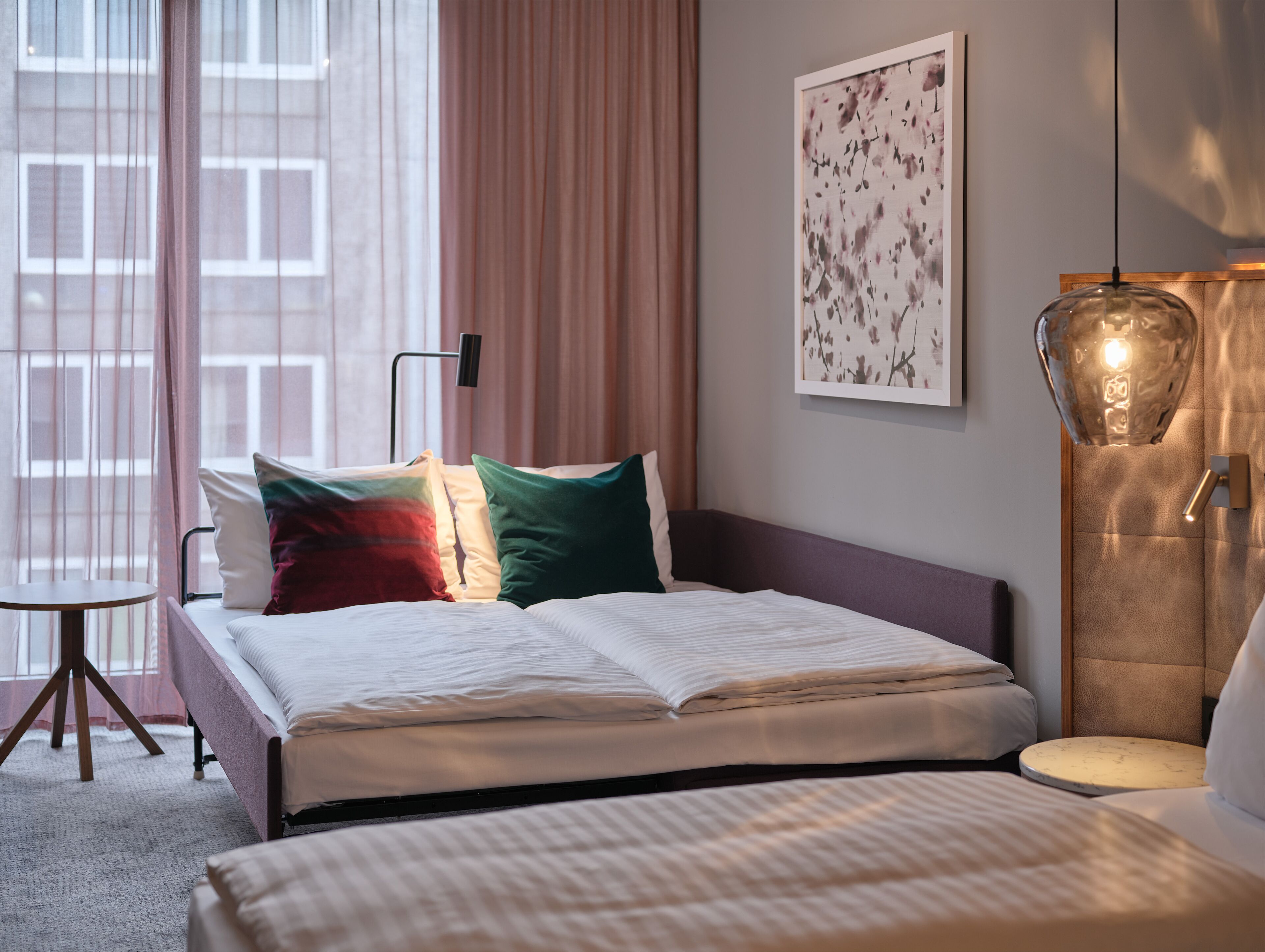 A hotel room with a bed, a bedside table, a lamp, and a window with curtains.
