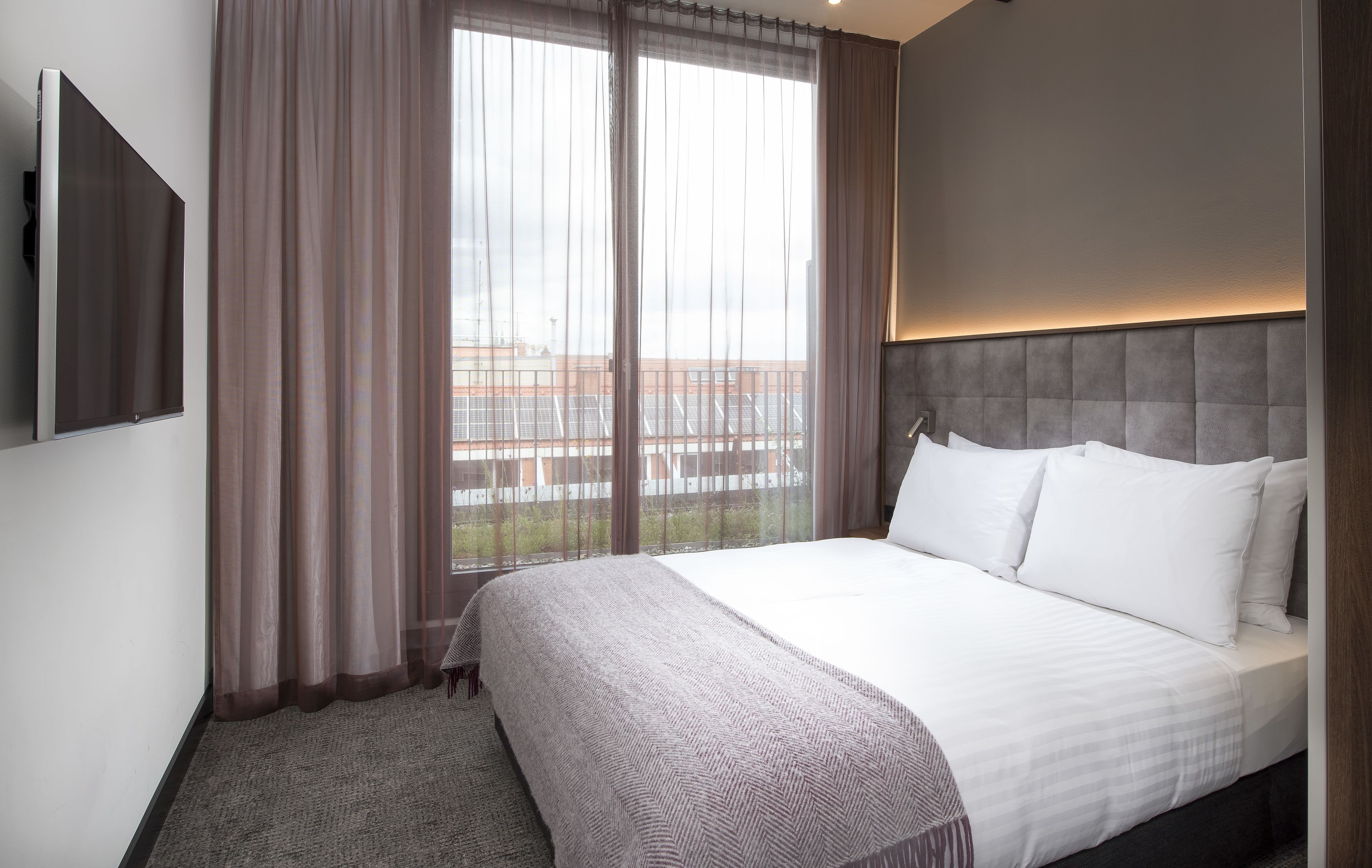 A hotel room with a large bed, a TV mounted on the wall, and a view of a building through sheer curtains.
