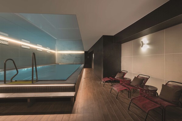 A modern indoor pool area with a clear pool, lounge chairs, and a wooden floor.