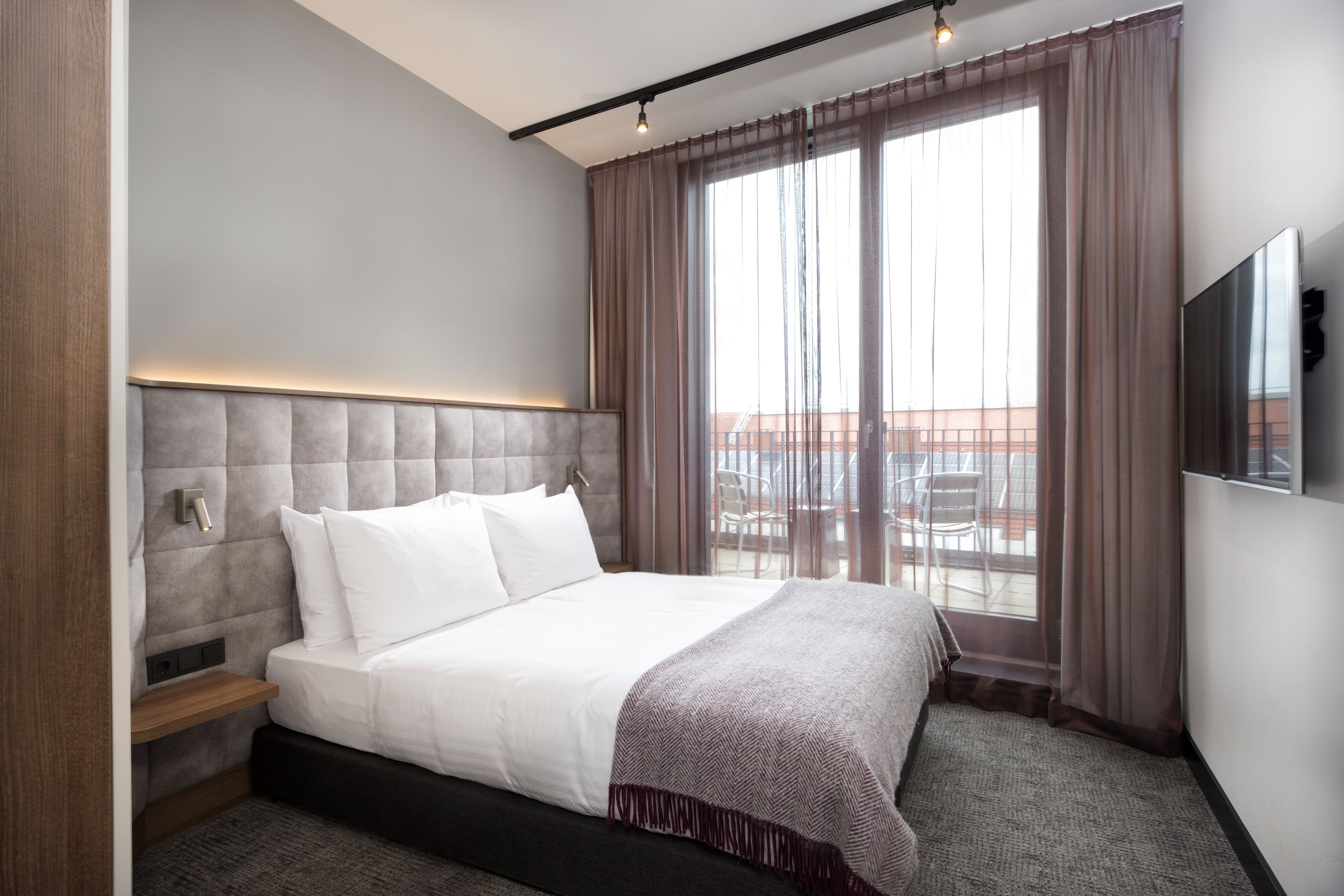 Une chambre d’hôtel moderne dotée d’un grand lit, d’un téléviseur fixé au mur et offrant une vue sur l’extérieur grâce à de grandes fenêtres.
