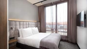 A modern hotel room with a large bed, a TV mounted on the wall, and a view of the outdoors through large windows.