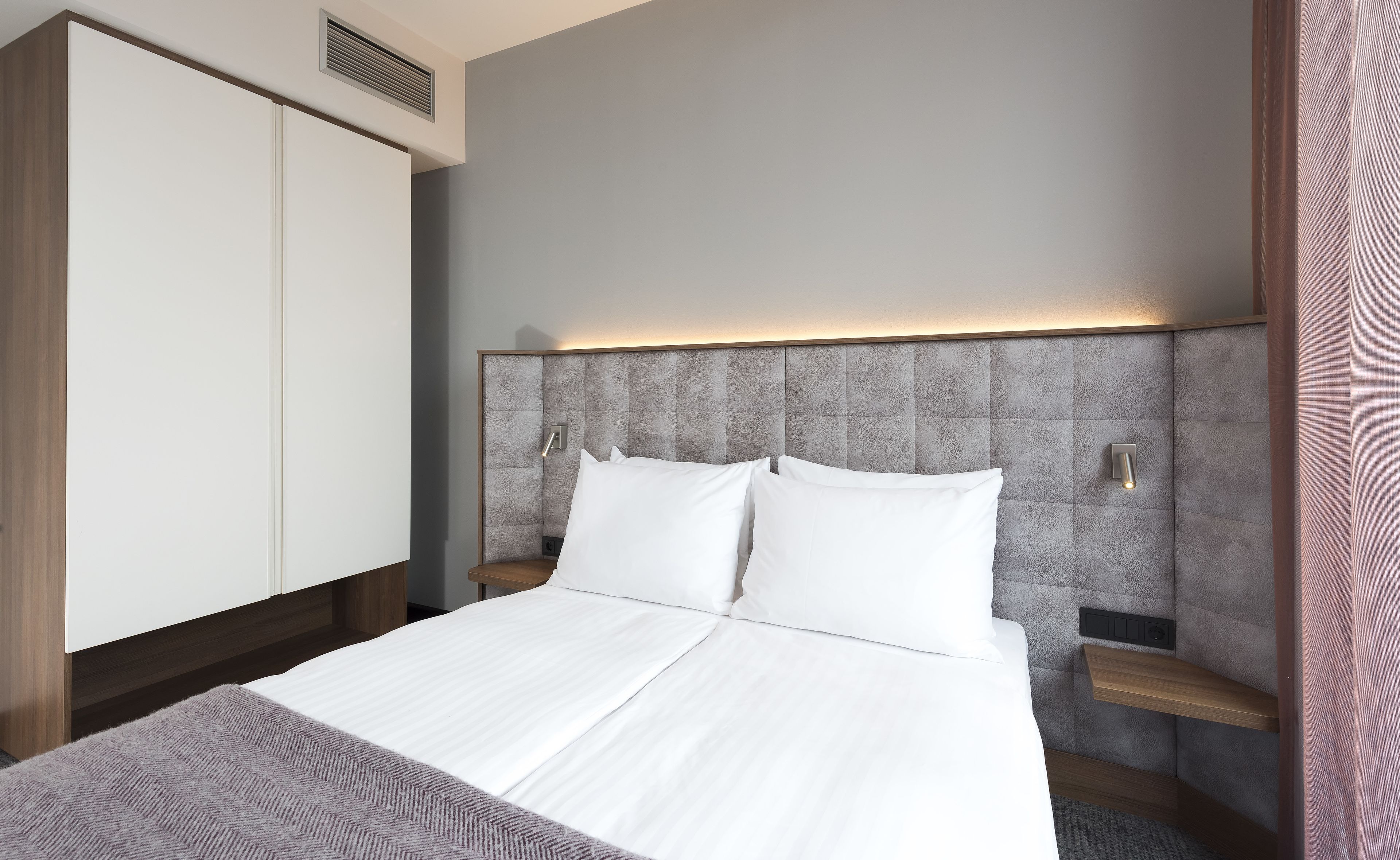 Una habitación de hotel moderna con una cama grande, un armario y un cabecero con iluminación integrada.