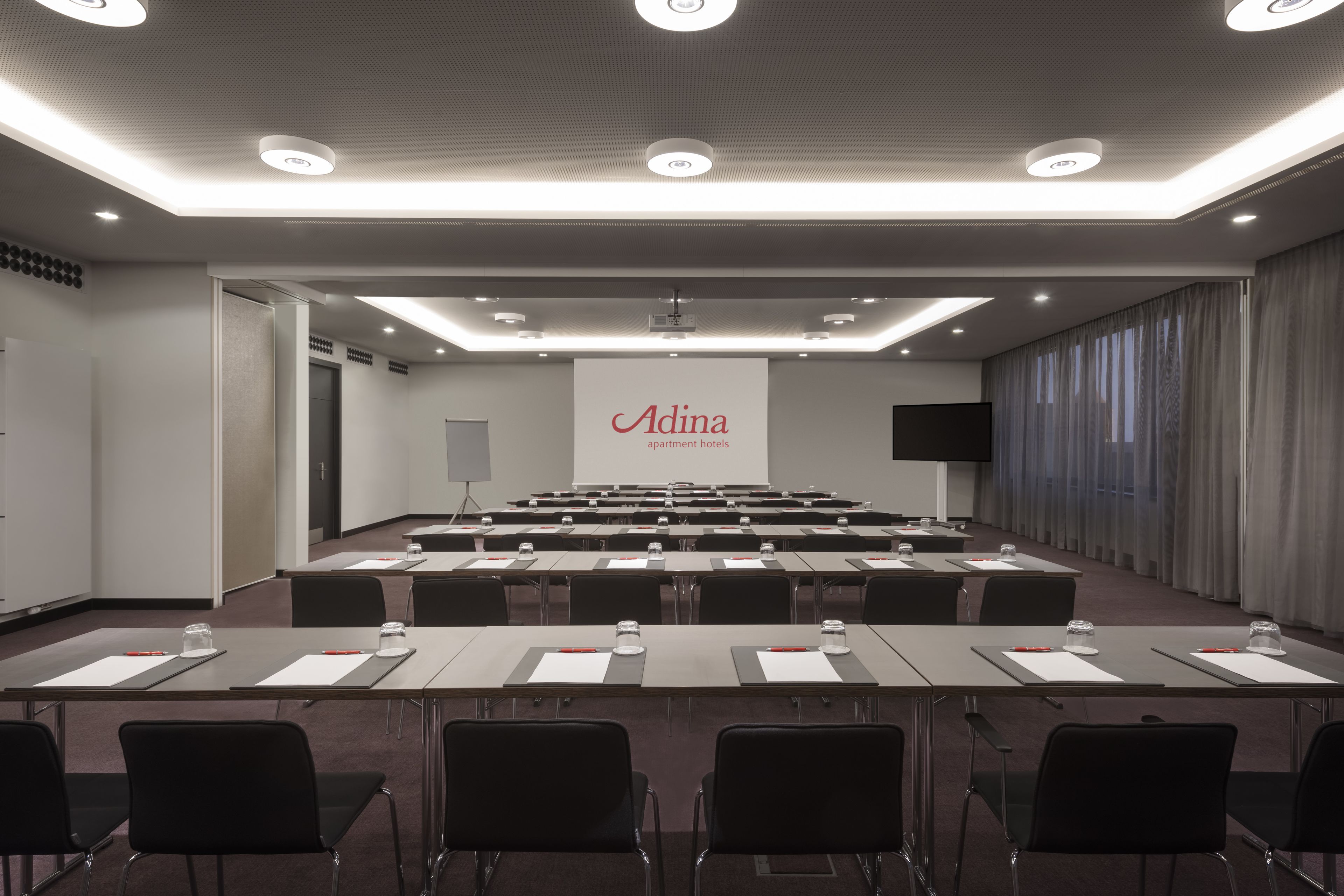 A conference room with a large screen displaying the Adina logo, multiple tables with chairs, and a projector.