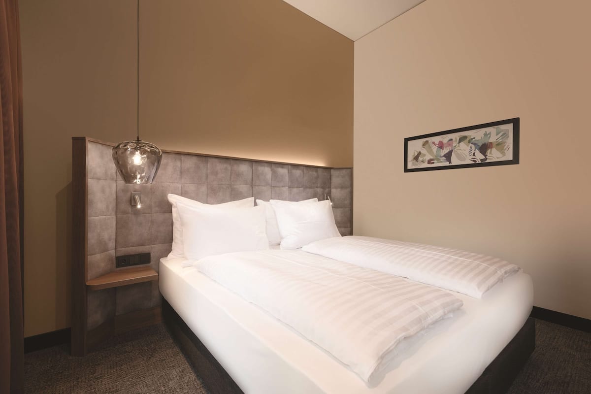Une chambre d’hôtel avec un grand lit, une tête de lit dotée d’un luminaire, une table de chevet et une œuvre d’art encadrée au mur.