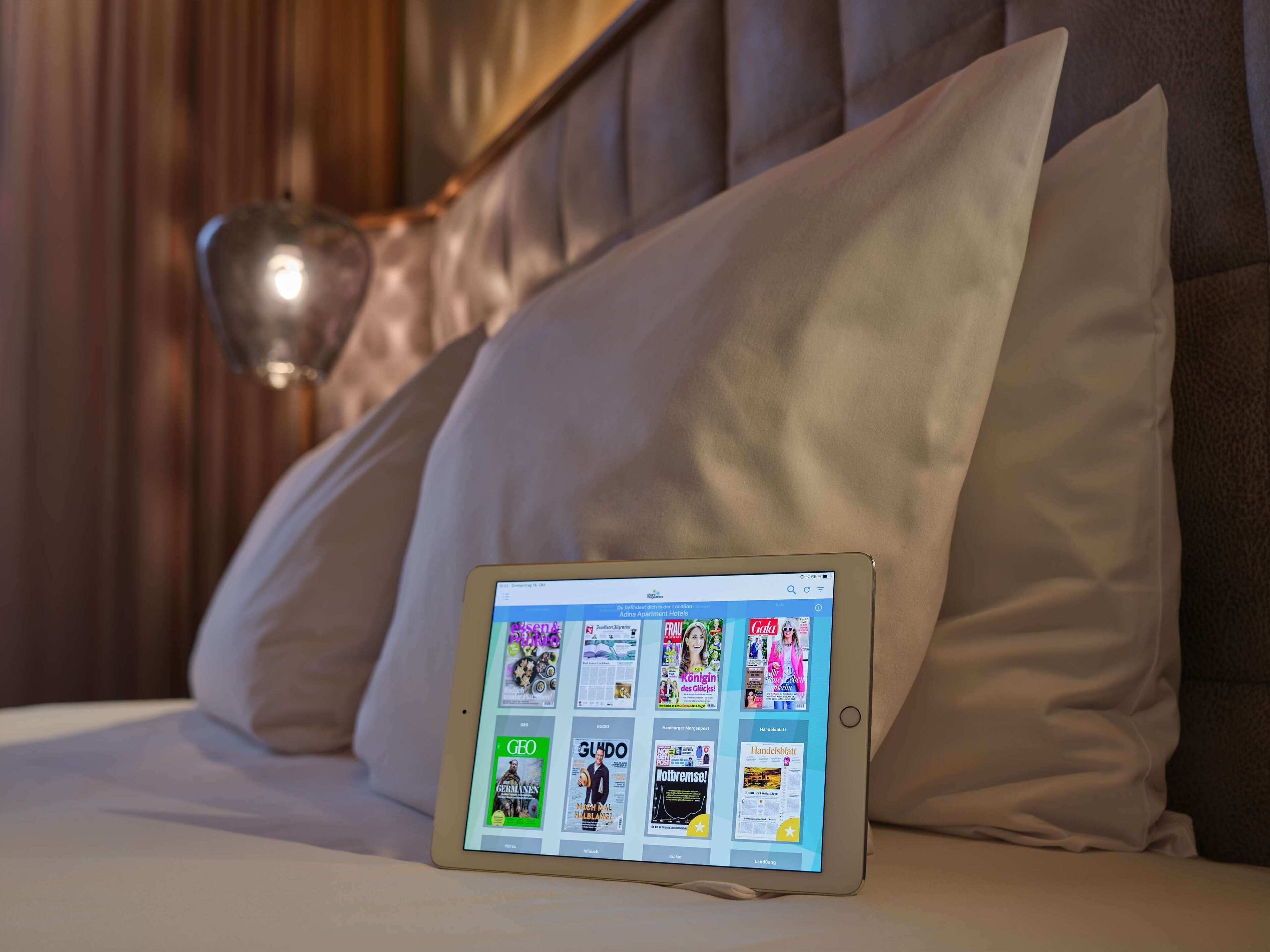 A tablet displaying a magazine layout on a bed with a large pillow and a lamp in the background.