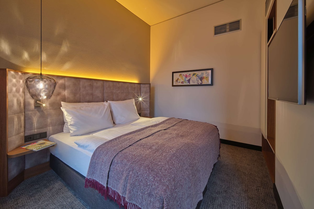 Une chambre d’hôtel avec un grand lit, une table de chevet avec une lampe, un tableau encadré au mur et un téléviseur fixé au mur.