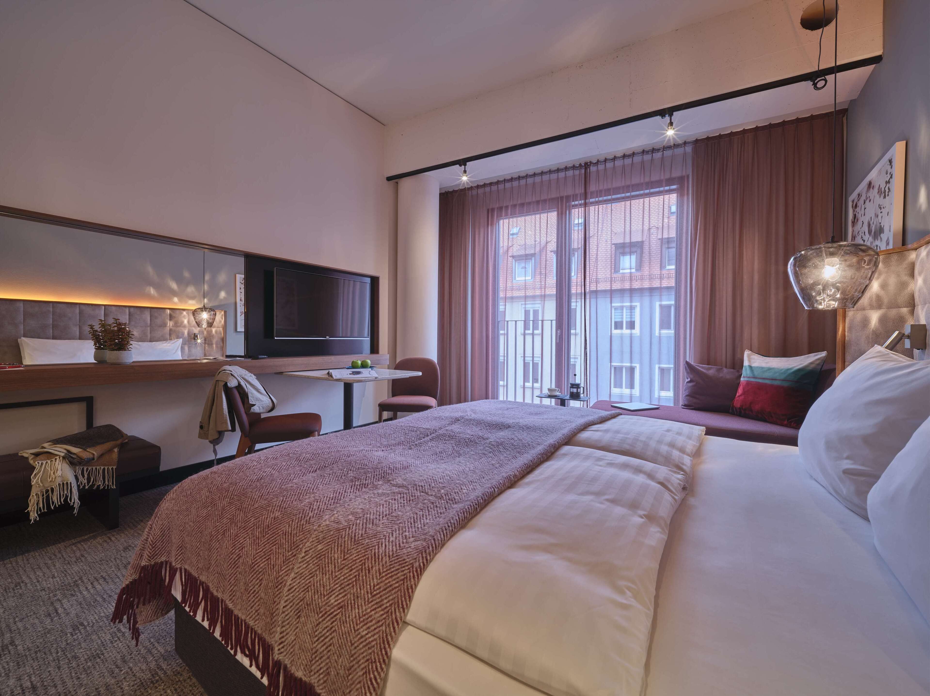 A hotel room with a bed, a desk with a TV, a chair, and a window with curtains.