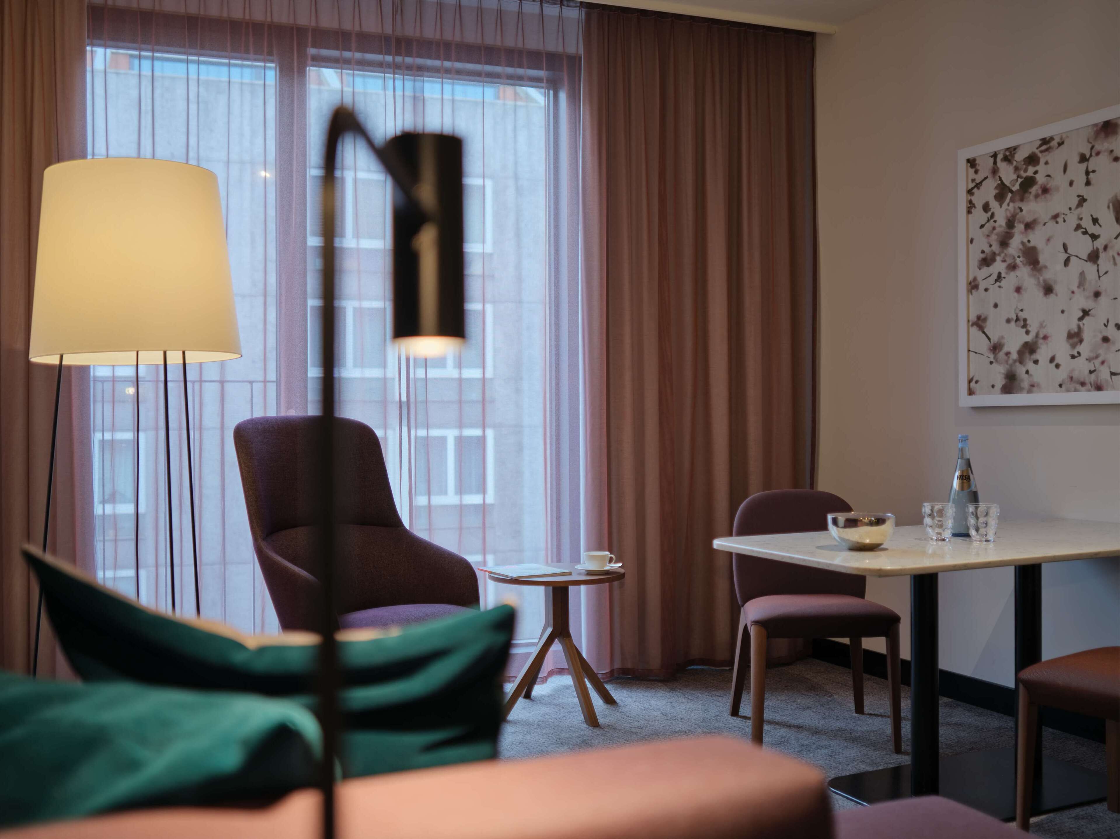 A hotel room with a large window, a table with a bottle and glasses, a chair, and a floor lamp.