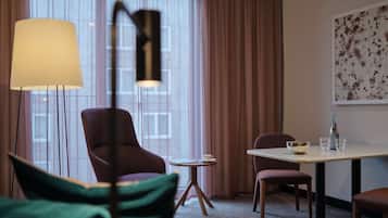 A hotel room with a large window, a table with a bottle and glasses, a chair, and a floor lamp.