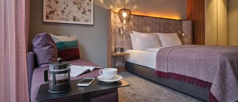 A modern hotel room with a bed, a bedside table with a lamp, a coffee maker, a cup, and a smartphone on a small table.
