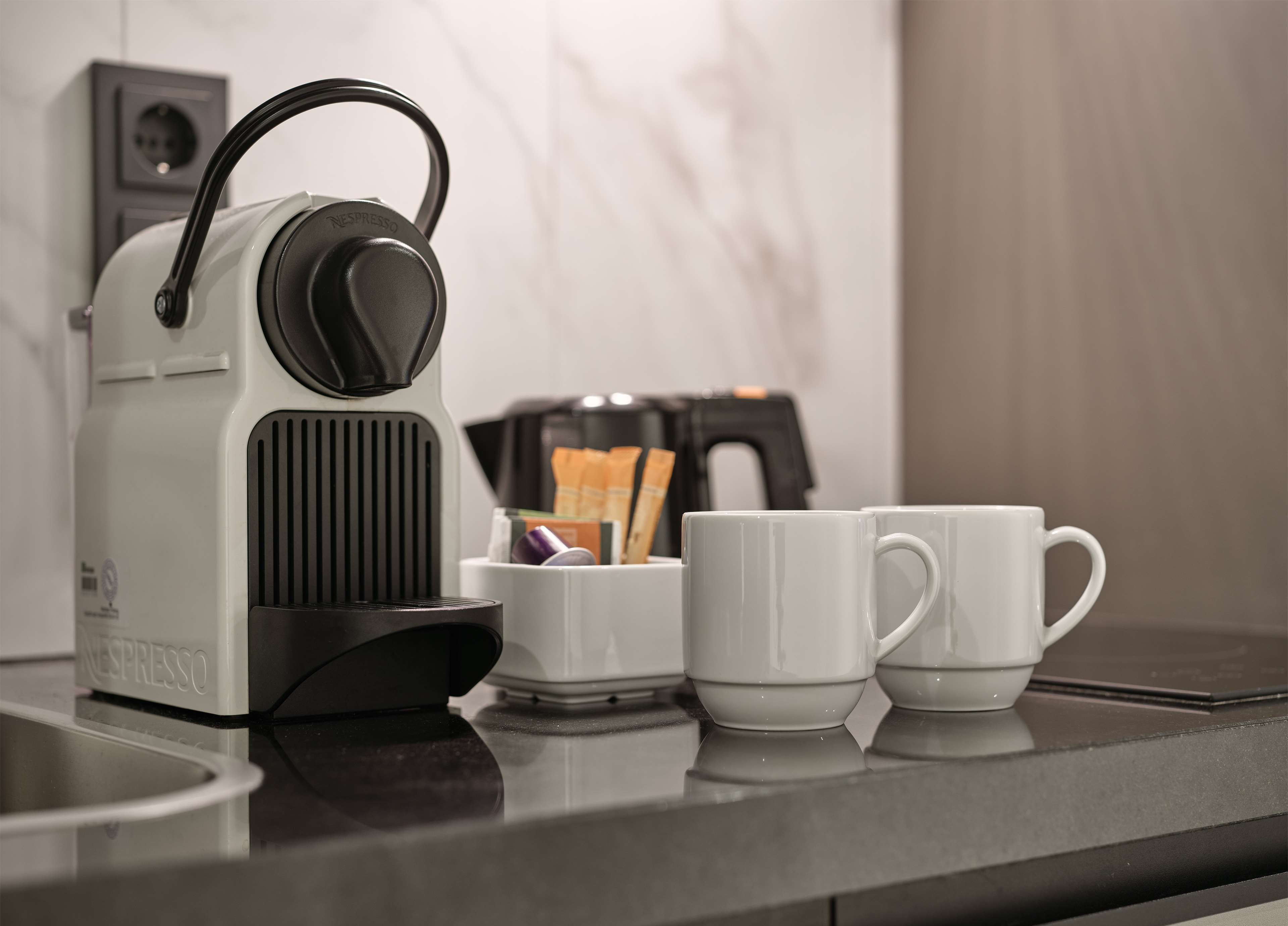 Een Nespresso koffieapparaat op een aanrecht, met twee witte kopjes en een klein bakje koffiekapsules.