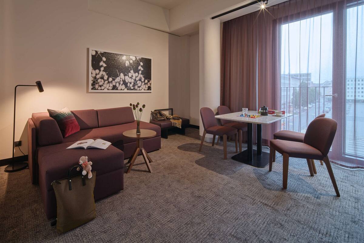 Une chambre d’hôtel moderne avec un canapé, une petite table ronde, une table à manger avec des chaises, une grande fenêtre avec des rideaux et une vue sur la ville.