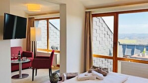 Habitación de hotel con cama, escritorio con silla, televisión y ventanas grandes con vistas.