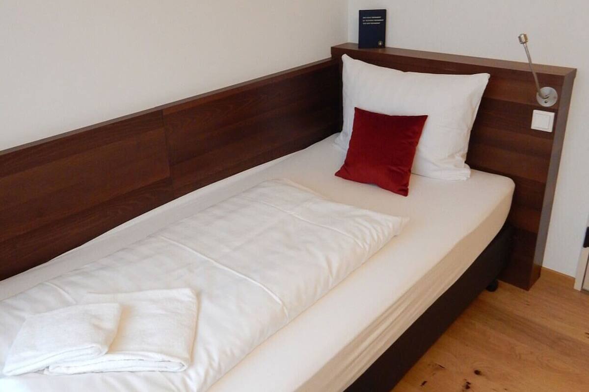 Een enkel bed met witte lakens, een rode kussen en een houten hoofdbord in een kamer met een houten vloer en een klein tapijt.