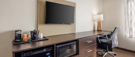 A hotel room with a flat-screen TV mounted on the wall, a microwave, a coffee maker, and a desk with a chair.