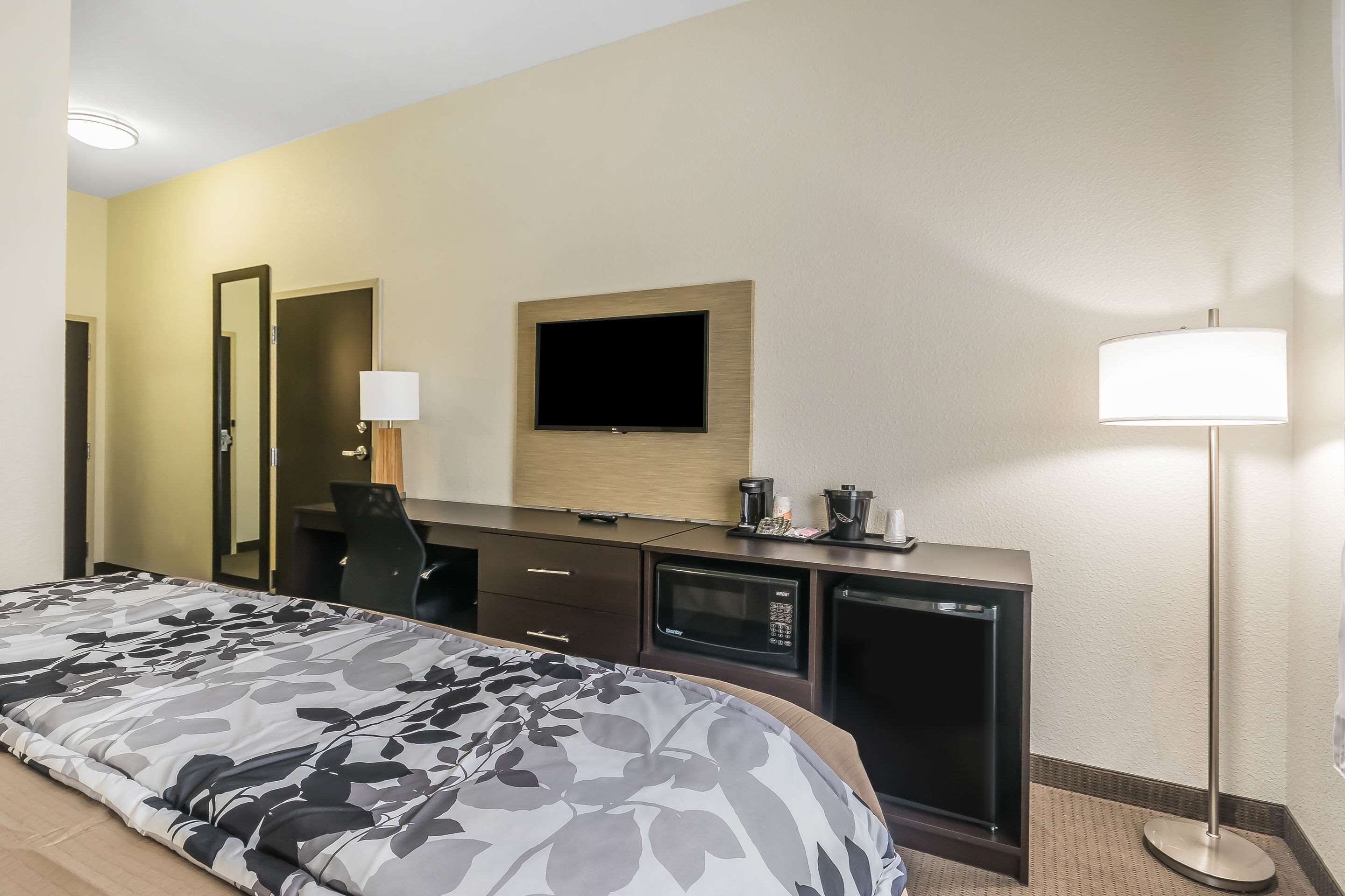 A hotel room with a bed, a desk with a flat-screen TV, a microwave, a coffee maker, and a lamp.