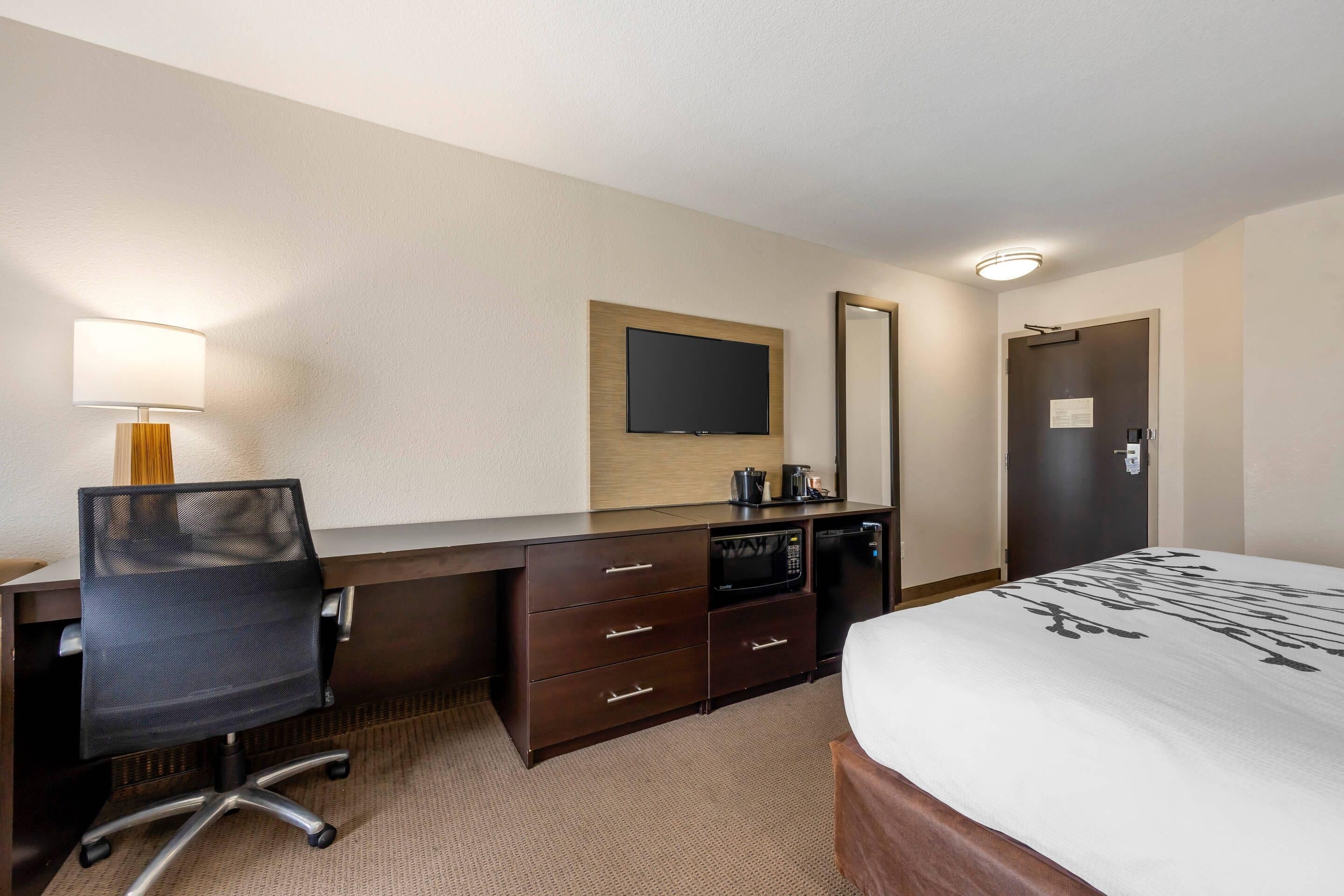 Habitación de hotel con cama, escritorio y silla, televisor de pantalla plana, microondas, cafetera y una puerta con un cartel.