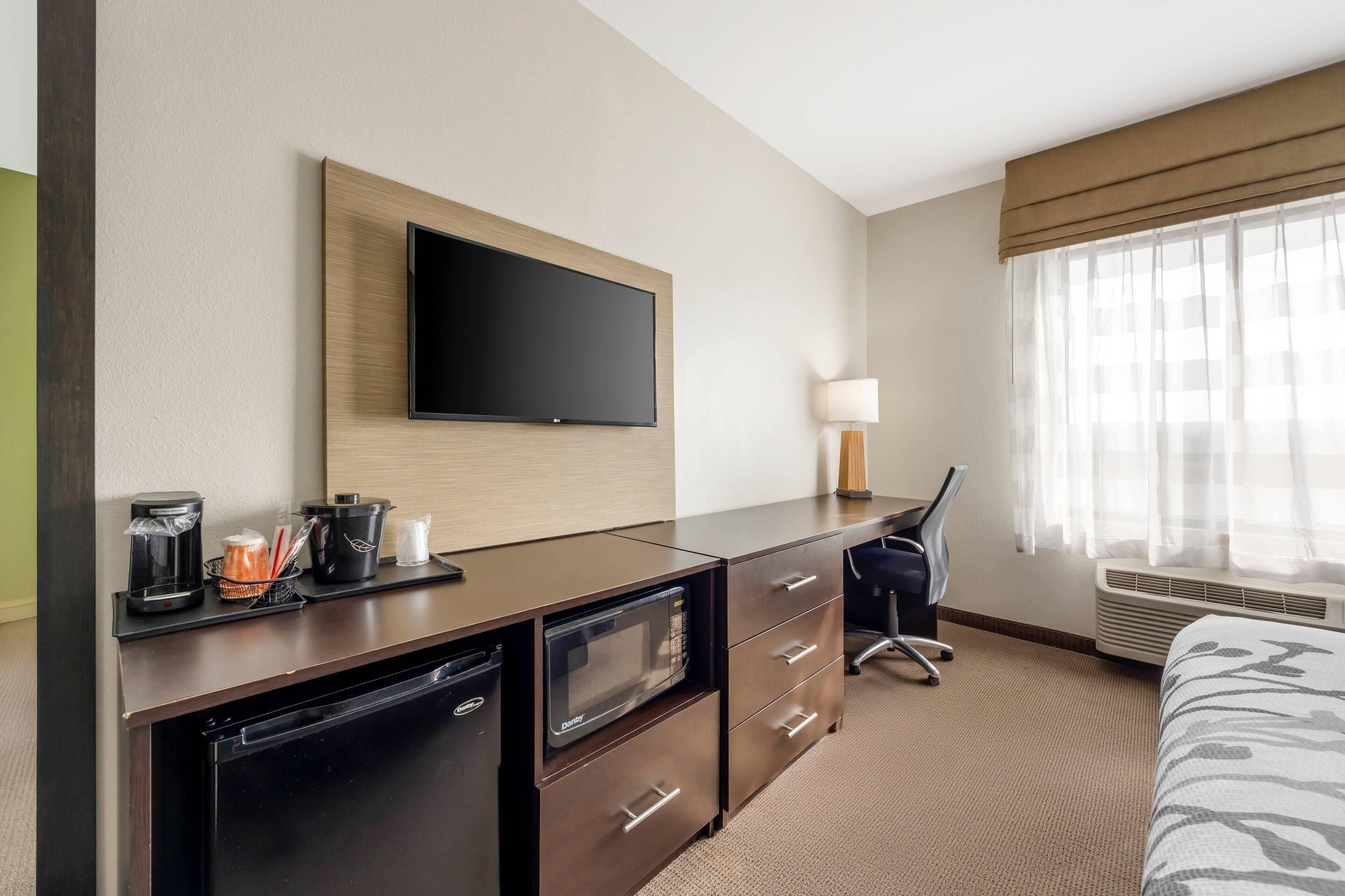 A hotel room with a flat-screen TV mounted on the wall, a desk with a microwave, a coffee maker, and a lamp. There is a bed with a patterned bedspread and a window with sheer curtains.