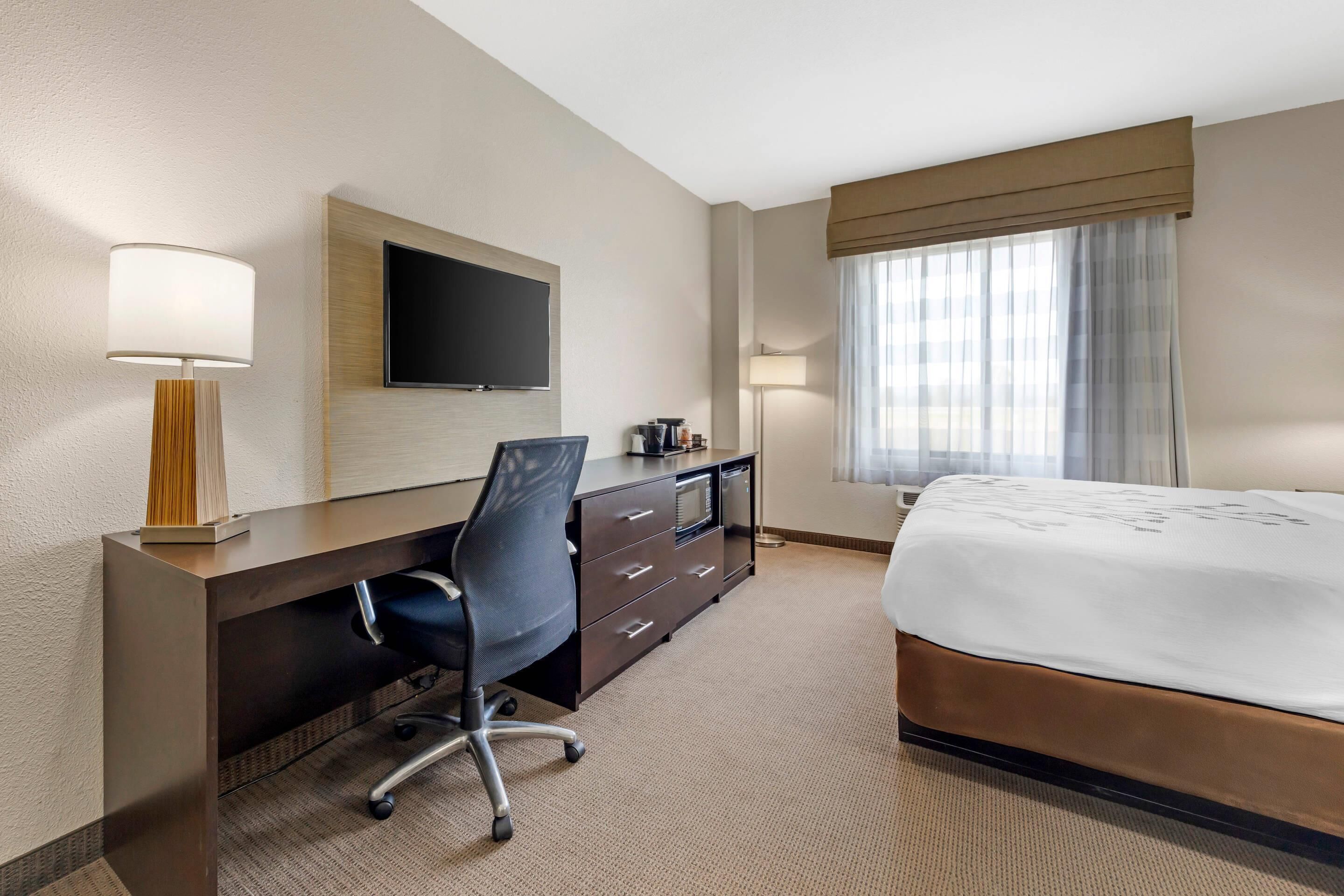 A hotel room with a bed, a desk with a chair, a television, and a window with curtains.