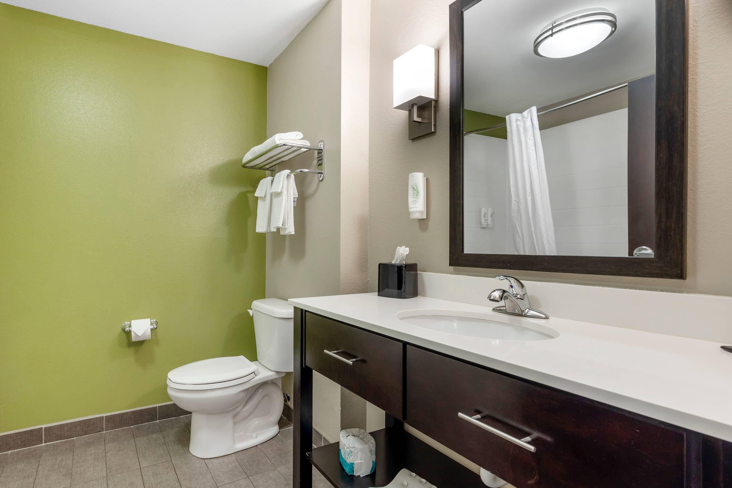 Un baño con pared verde, inodoro blanco y tocador con espejo y lavabo.