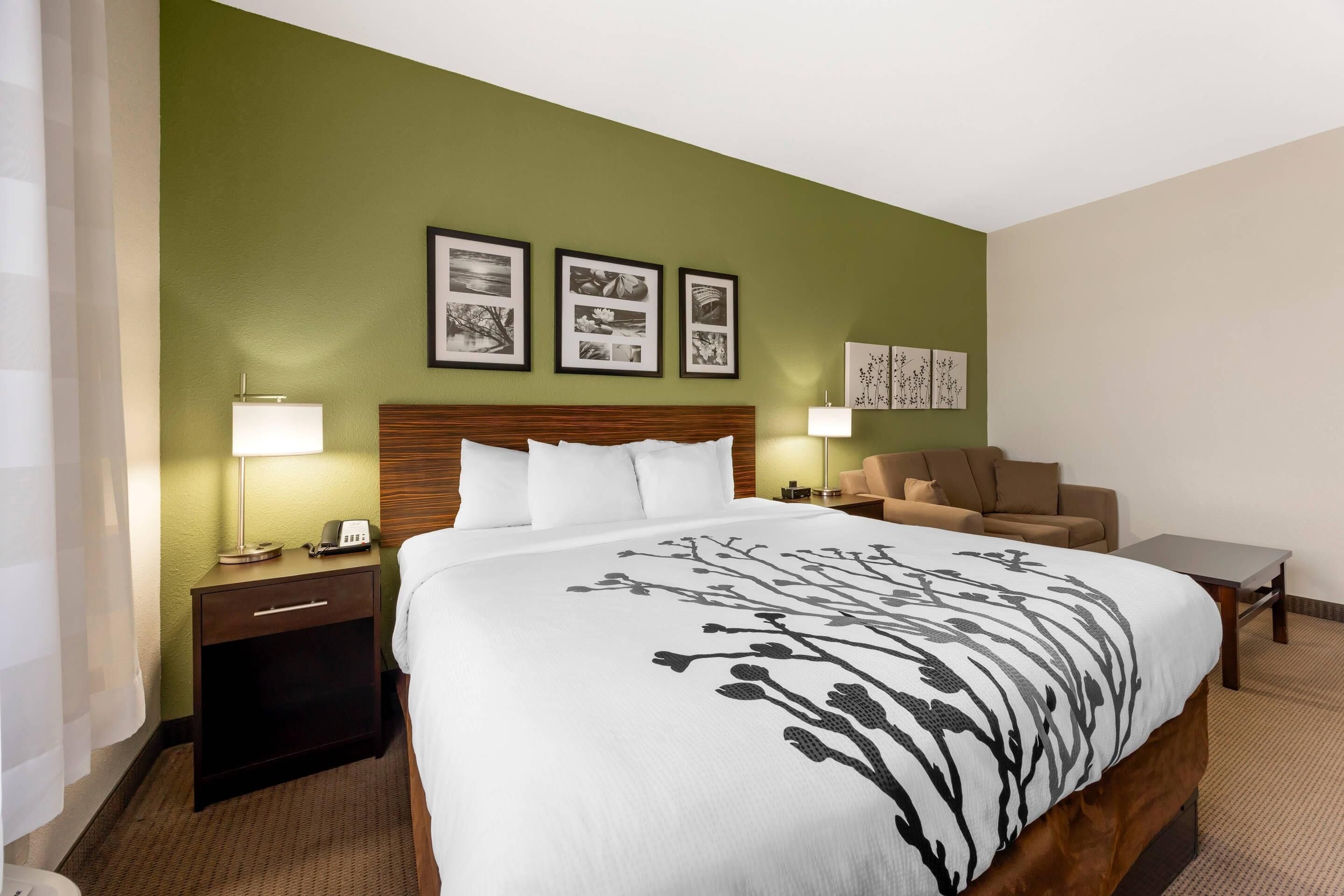 Habitación de hotel con una cama grande, cabecera de madera, mesita de noche con lámpara, una pequeña mesa y un sofá.