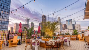 An outdoor seating area with chairs and tables, string lights, and umbrellas, overlooking a city skyline. - Freehand Los Angeles (Los Angeles)