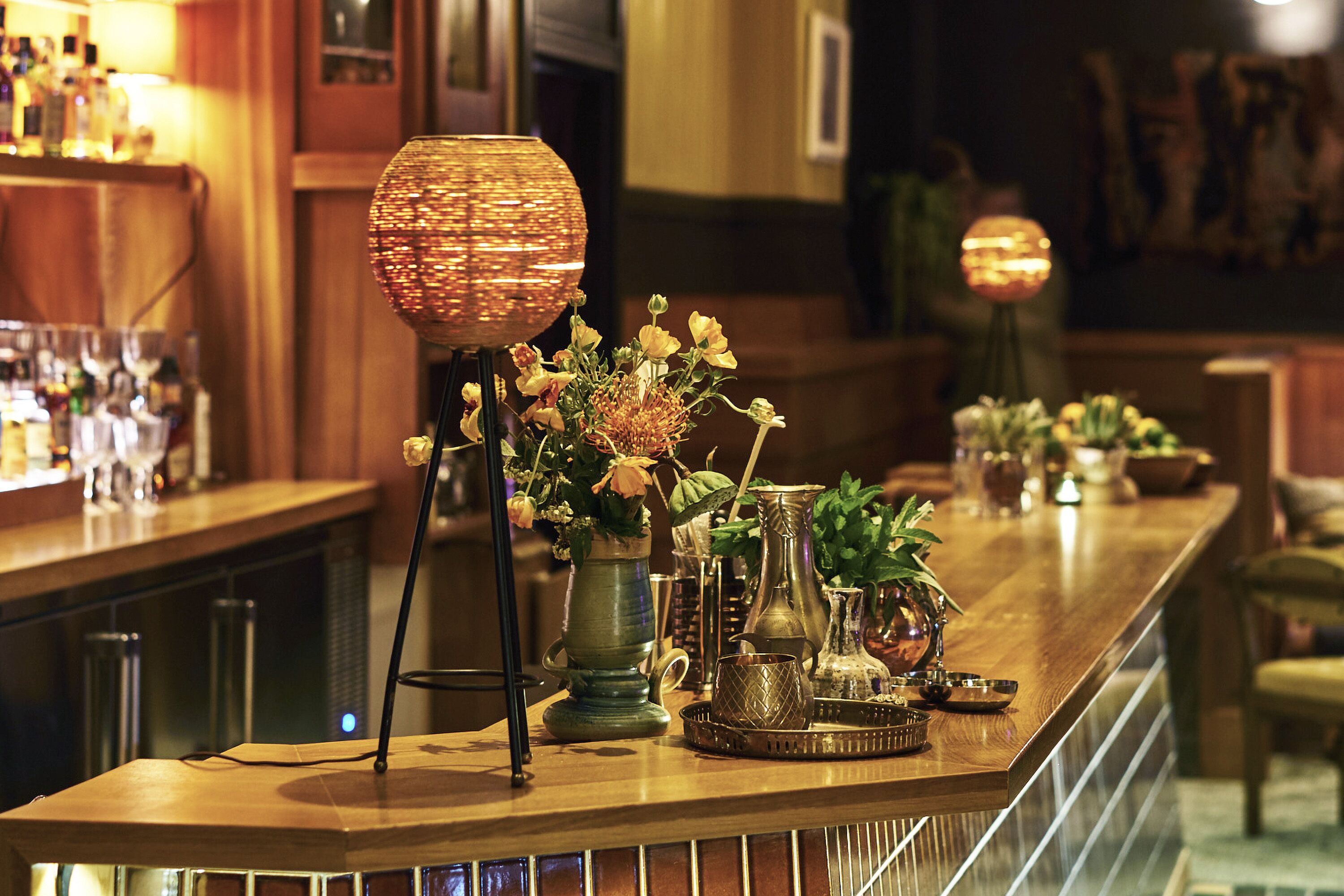 Una zona de bar con mostrador de madera, diferentes vasos y jarrones decorativos con flores.
