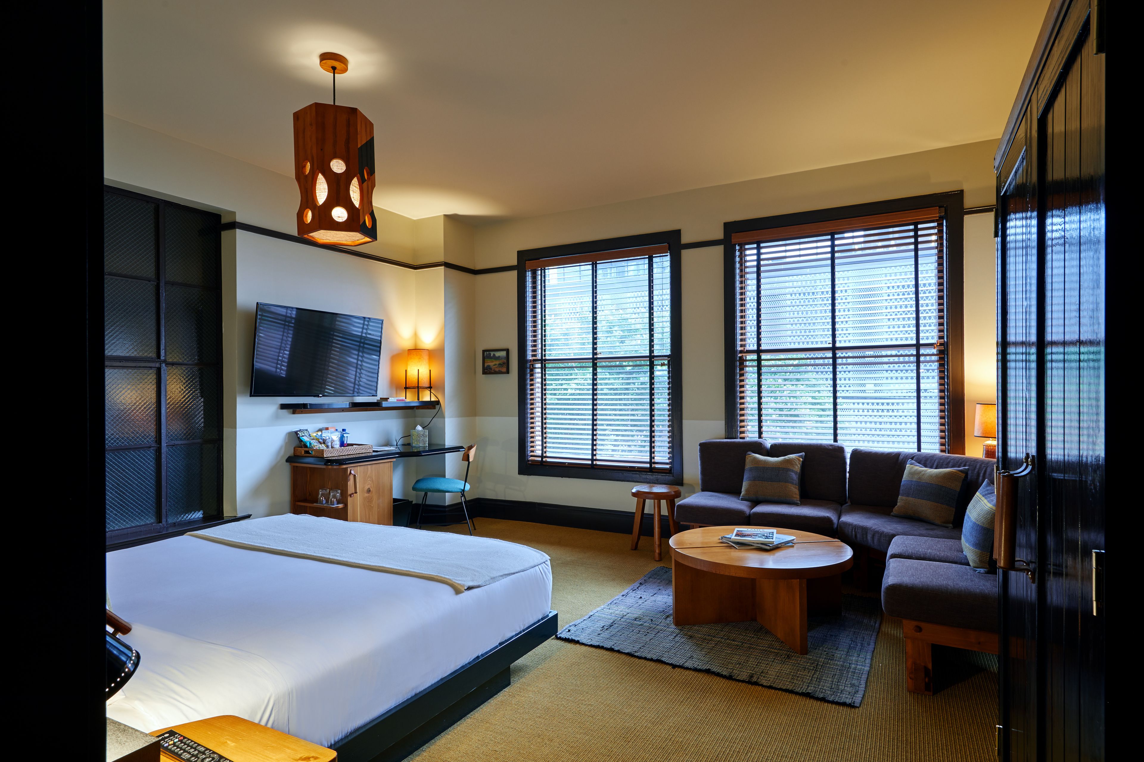 A hotel room with a bed, a sofa, a coffee table, a TV, and large windows with blinds.