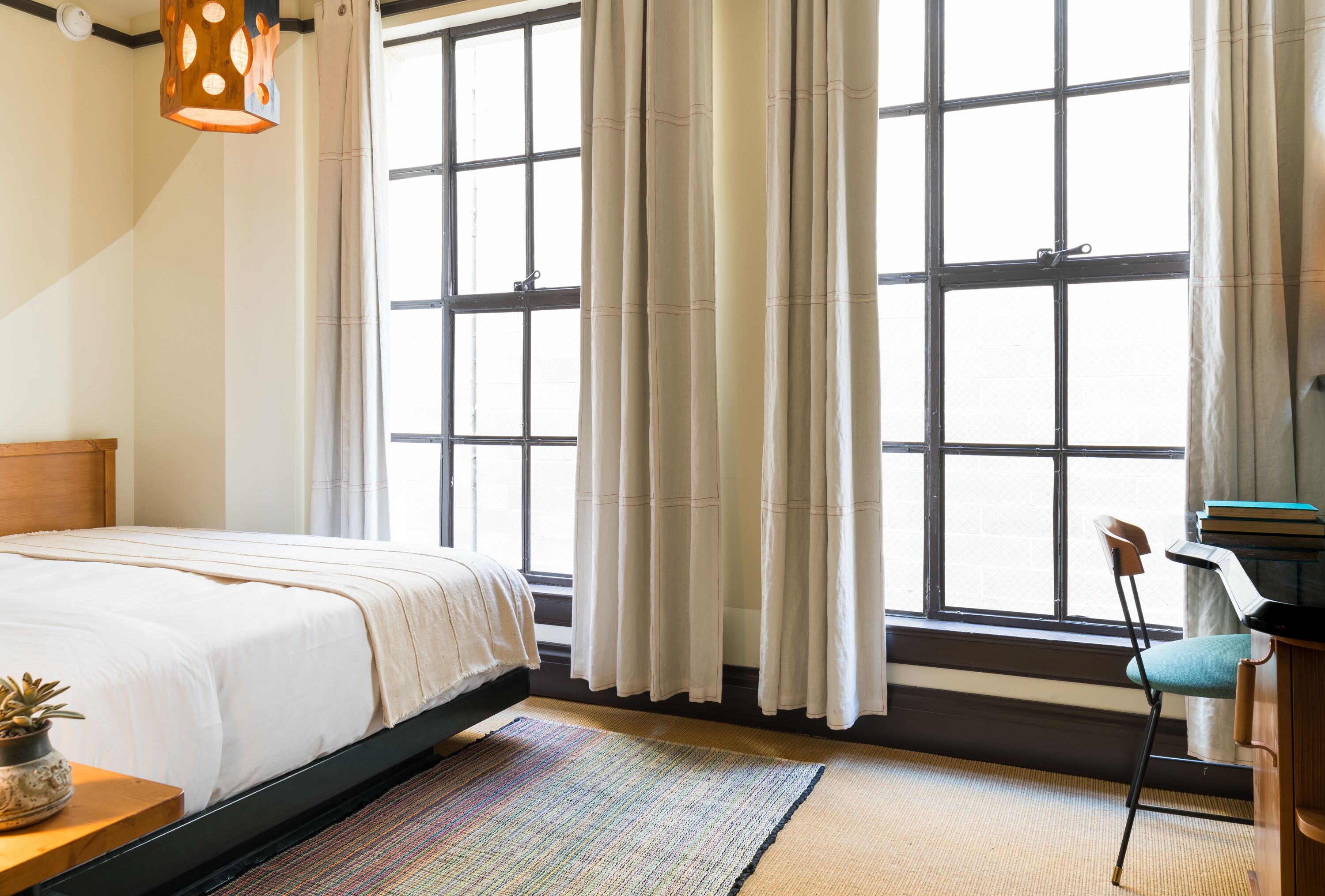 A bedroom with a bed, a desk, a chair, a window with curtains, and a hanging light fixture.