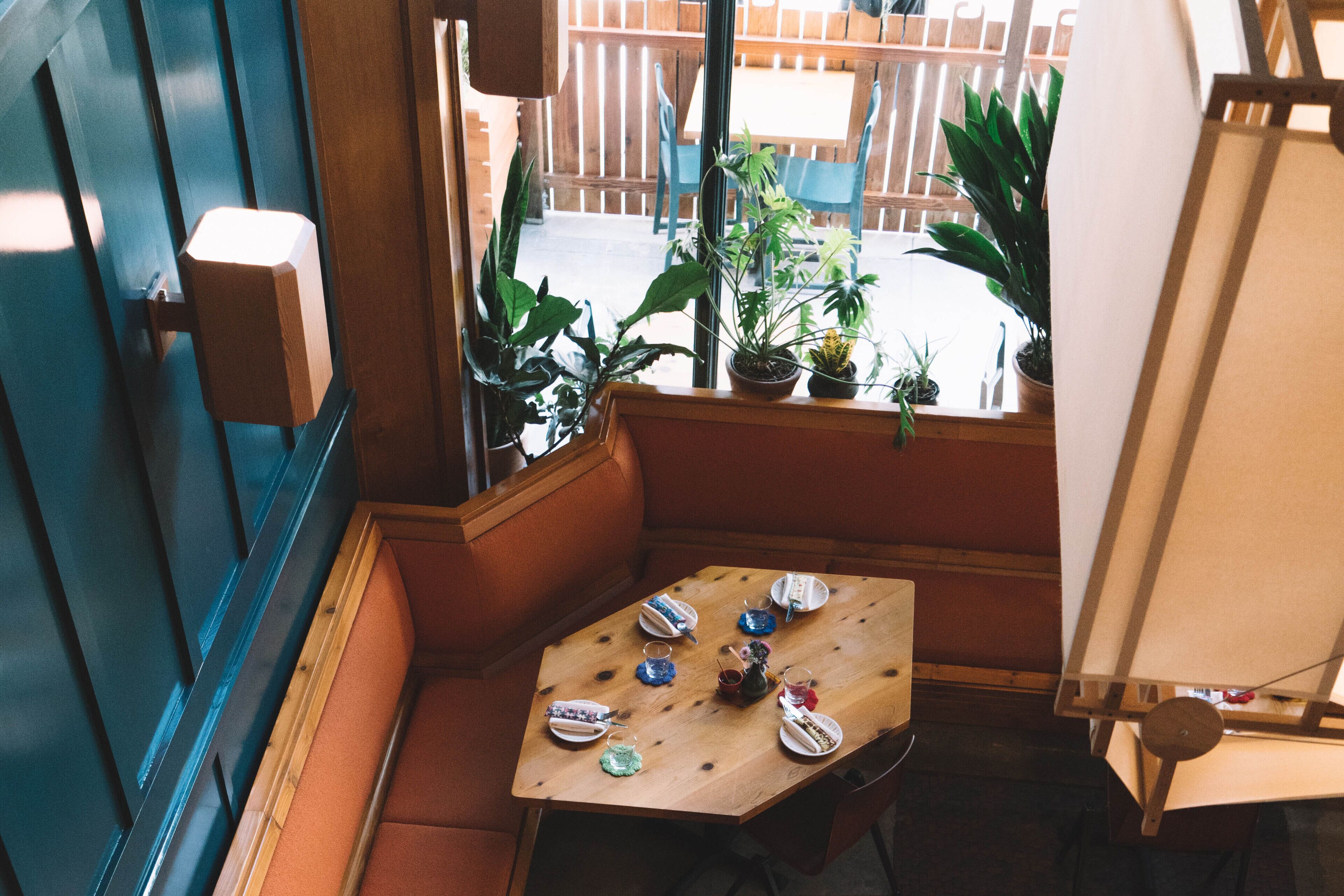 A corner seating area with a wooden table set for a meal, surrounded by potted plants and a view of the outside.