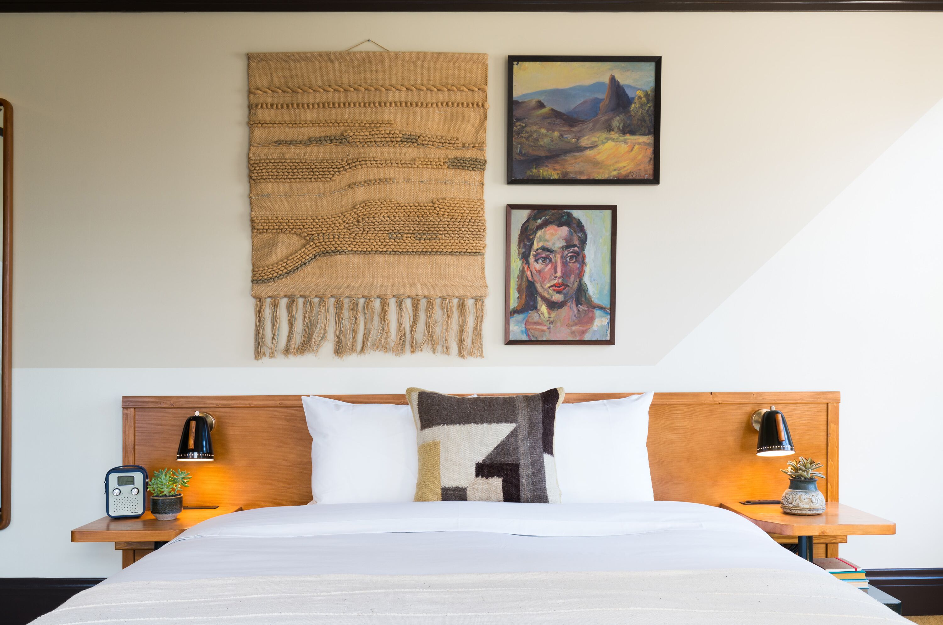 Una cama bien hecha con edredón blanco, un cojín estampado con motivos geométricos y un tapiz tejido sobre ella. Hay dos mesitas de noche con lámparas, una radio y una pequeña planta en maceta.