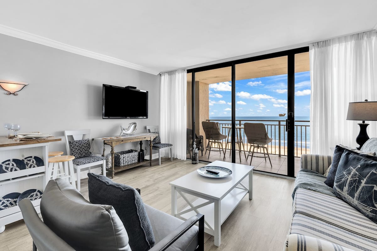 A modern hotel room with a balcony, a flat-screen TV, a dining area, and a sofa.