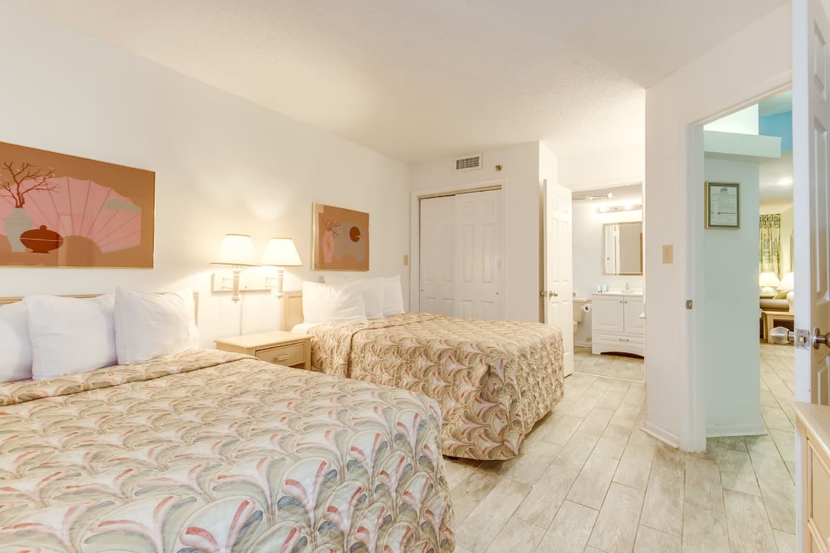 A hotel room with two beds, a nightstand, a lamp, a door, and a bathroom visible through an open door.
