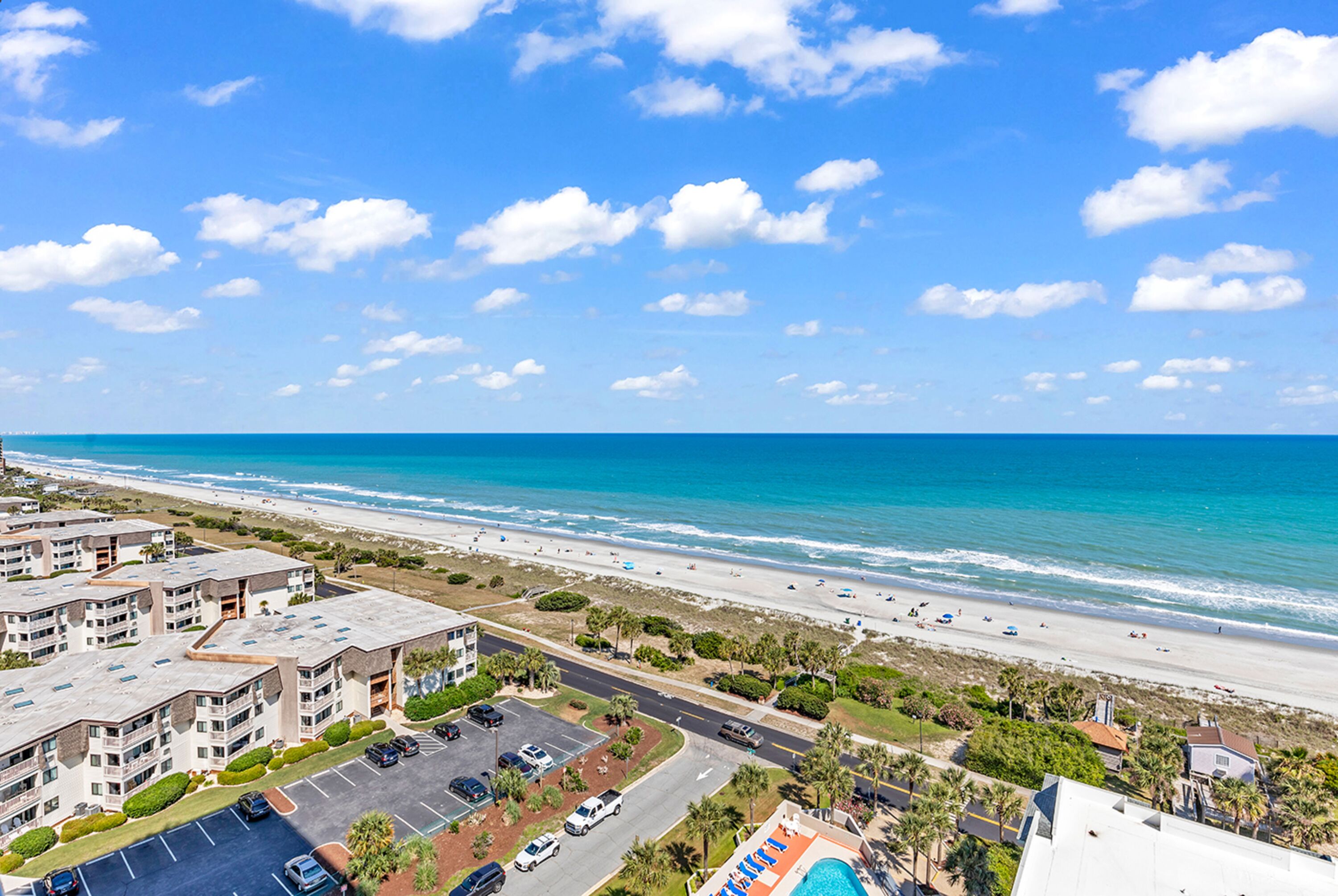 A coastal hotel with multiple buildings, a parking lot, and a beach view.