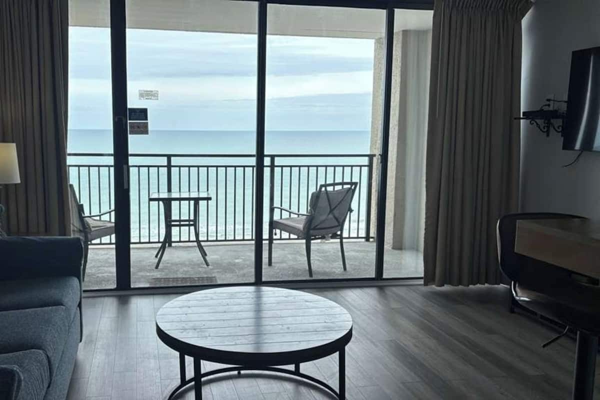 A hotel room with a balcony, a round table, a sofa, and a TV.