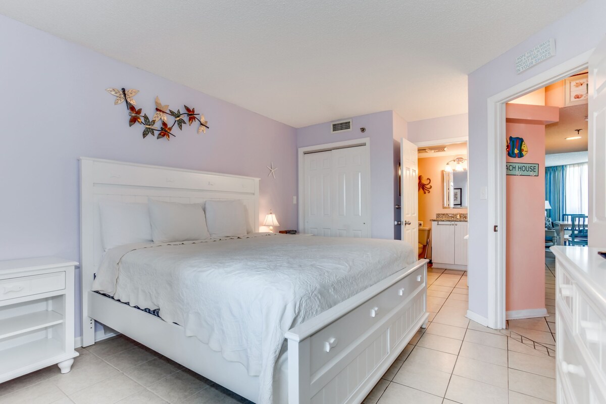 A bedroom with a white bed, a nightstand, a dresser, and a door leading to a bathroom.