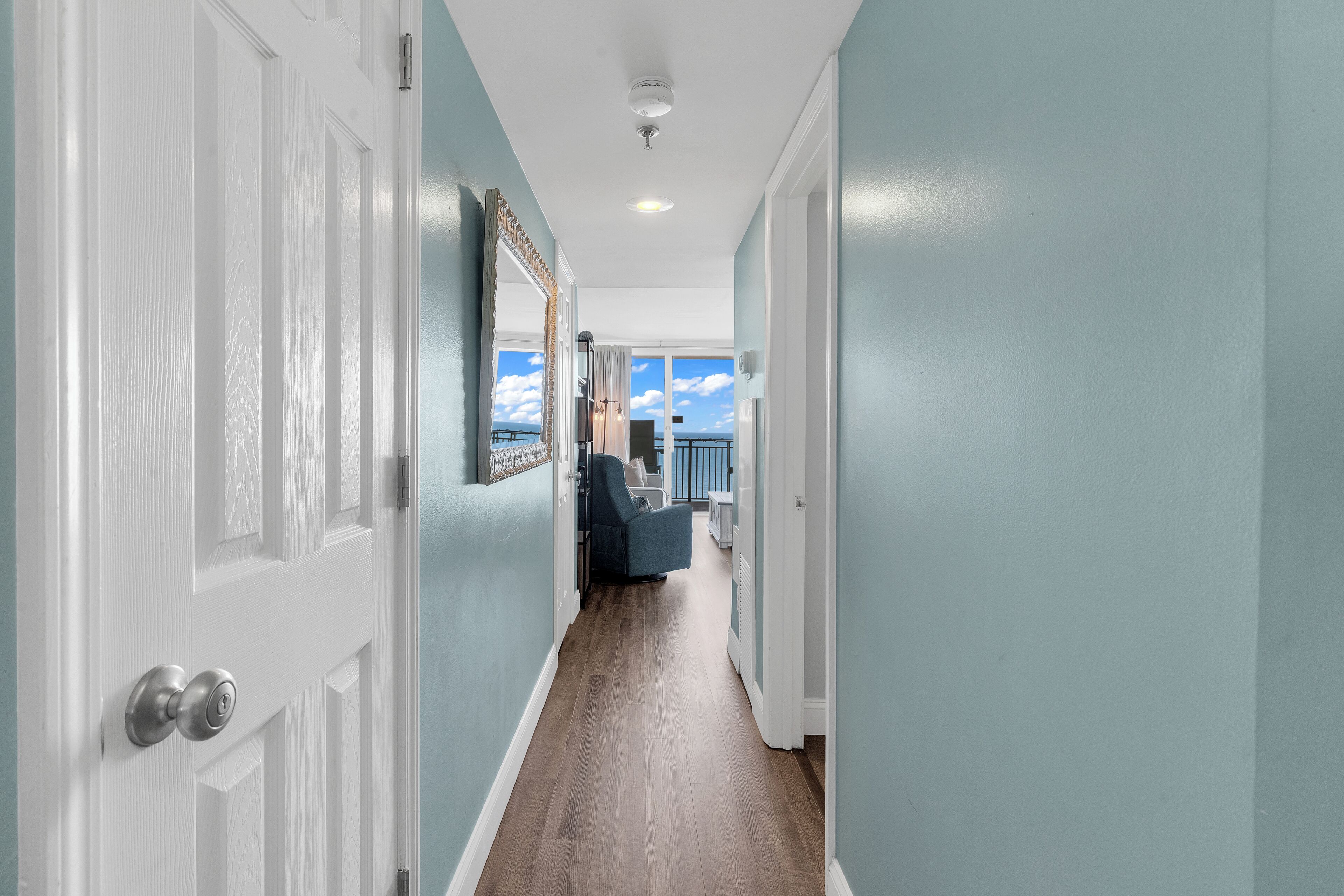 A hallway with a white door, a blue chair, and a view to the outside.