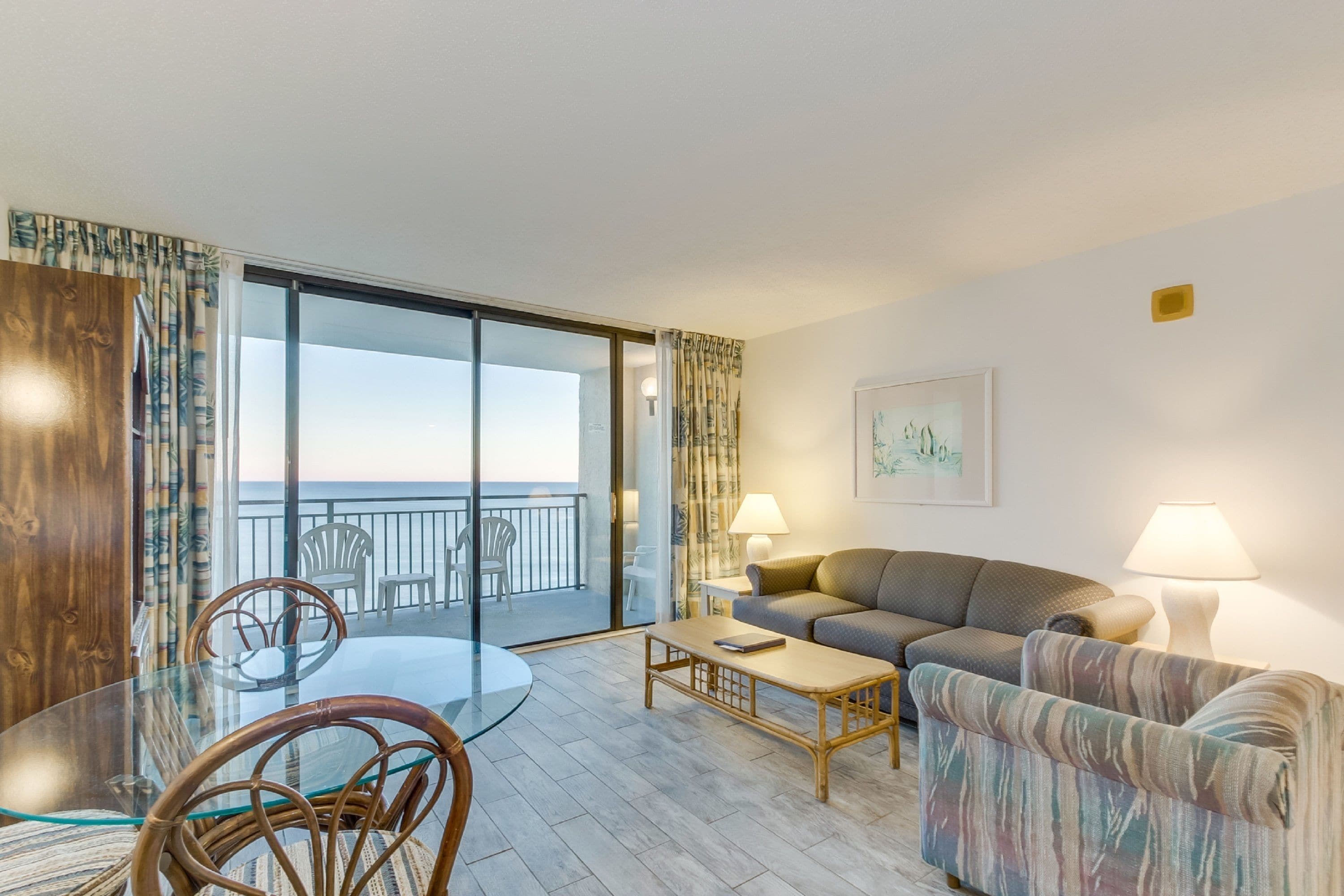A modern living room with a glass dining table, wicker chairs, a sofa, and a balcony with a view of the sea.