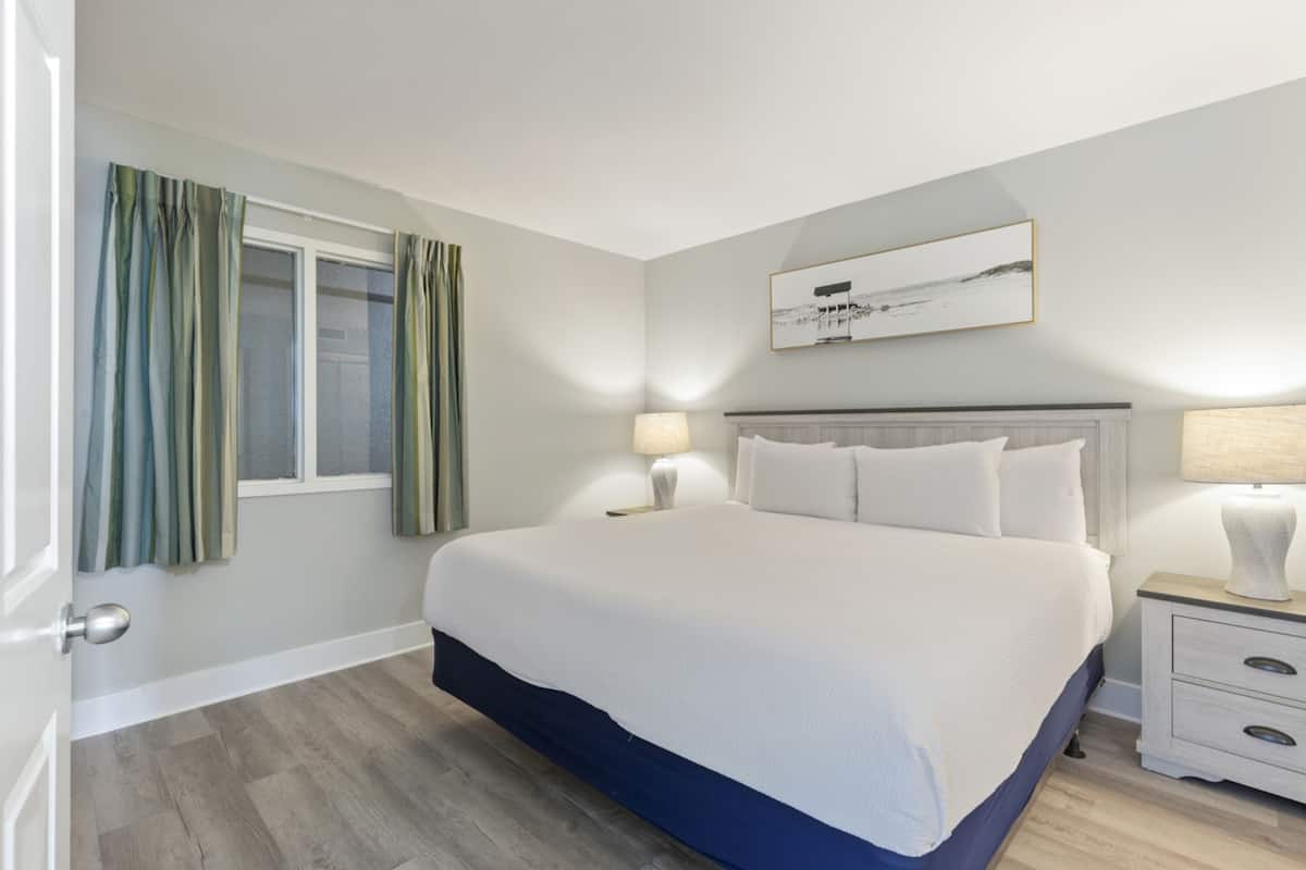A bedroom with a large bed, two bedside tables, a window with curtains, and a wall-mounted picture.