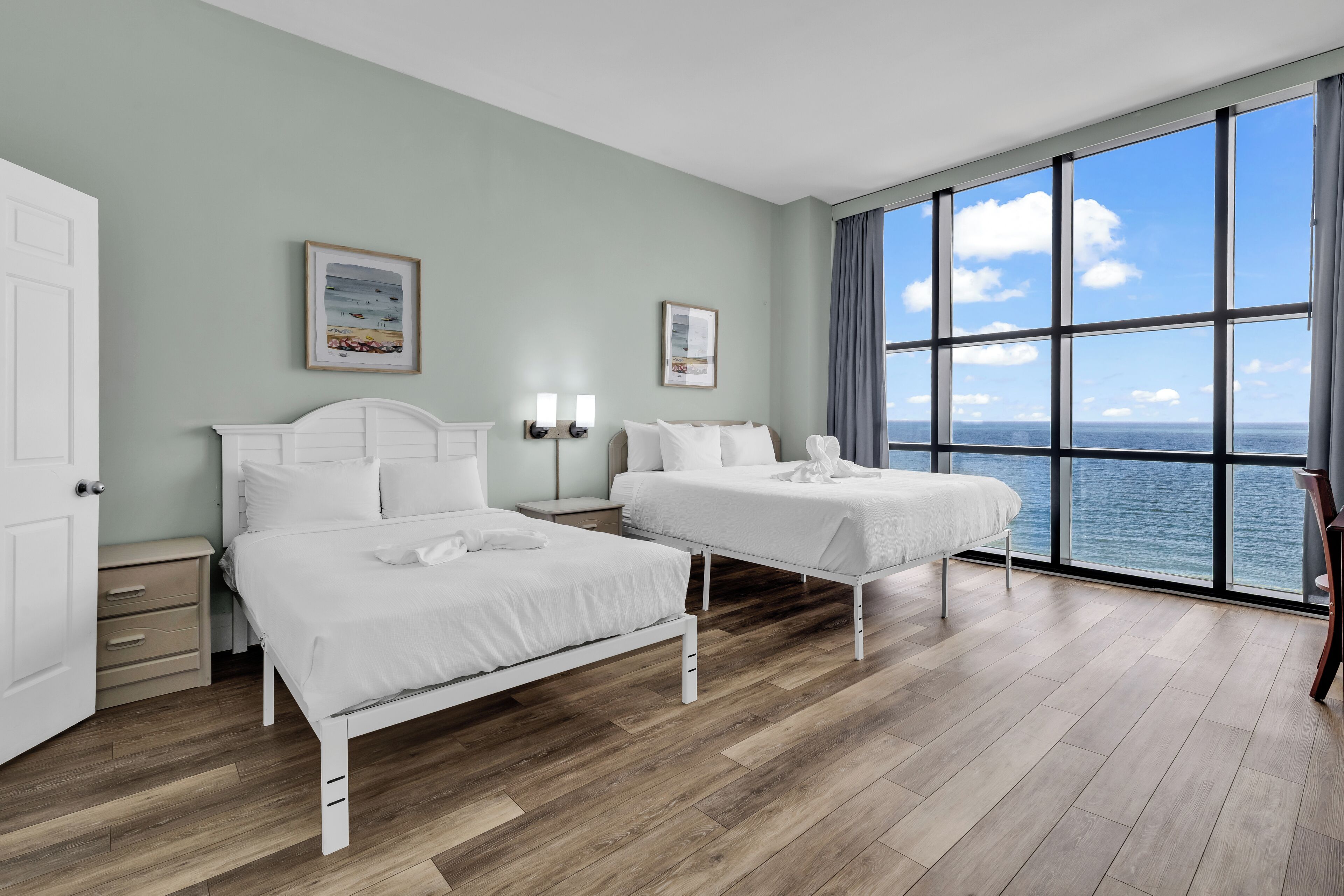 Habitación de hotel con dos camas, un ventanal con vista al mar, una puerta blanca y una mesita de noche con lámpara.