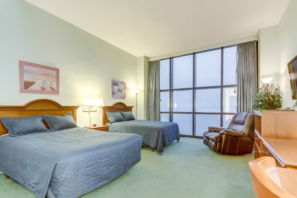 A hotel room with two beds, a desk, a chair, a lamp, and a large window.