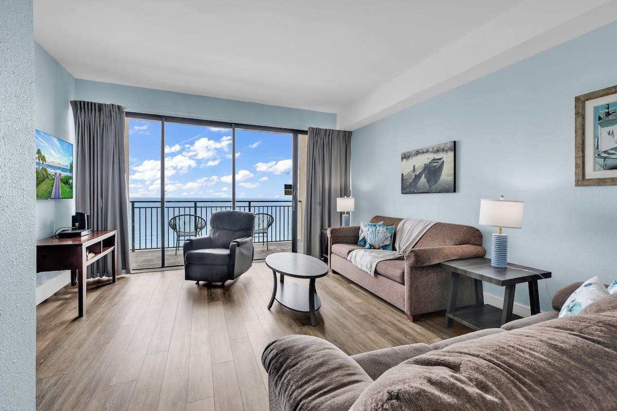 A hotel room with a balcony, a sofa, a chair, a small table, and a lamp.